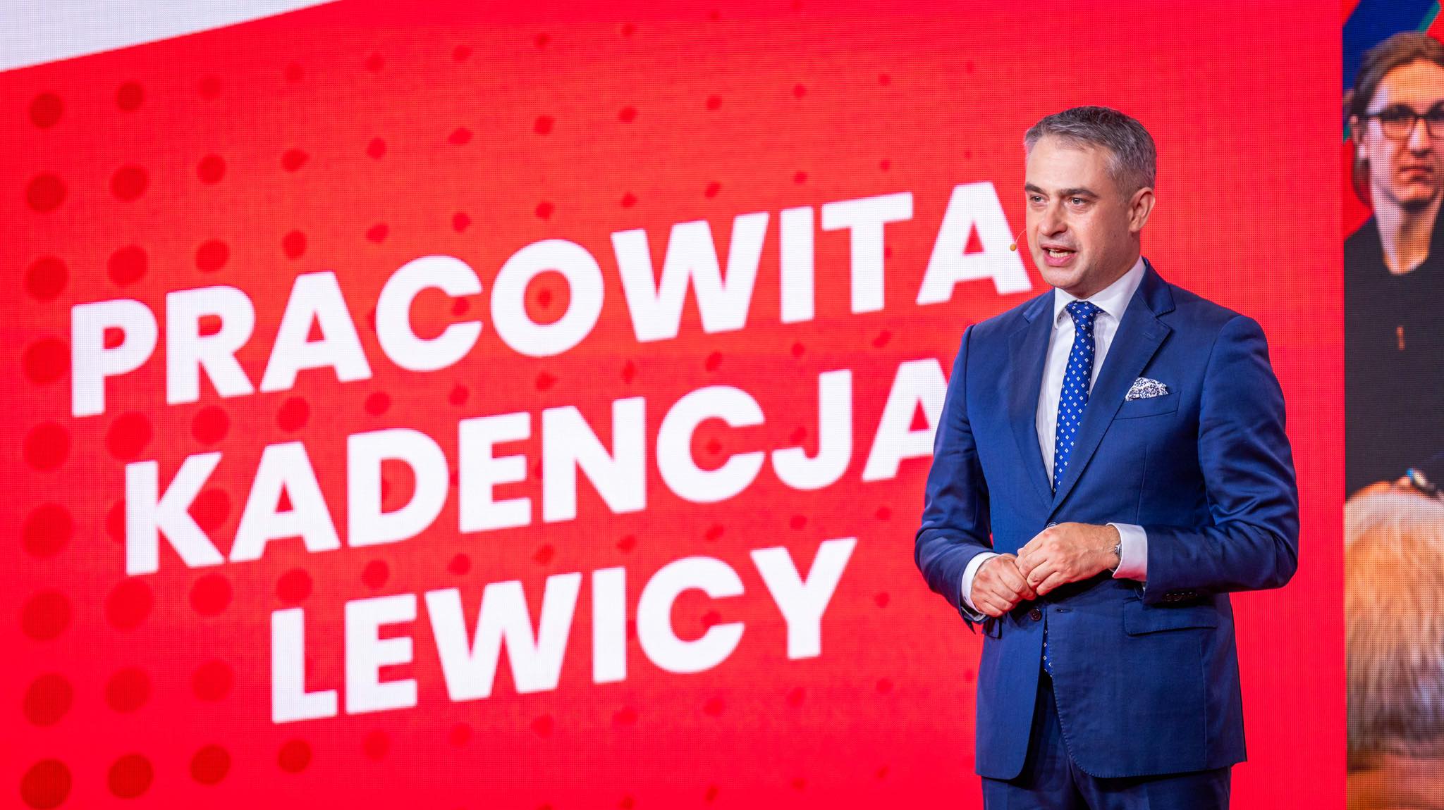 Mężczyzna w granatowym garniturze stoi na czerwonym tle z dużym białym napisem „PRACOWITA KADENCJA LEWICY”.
