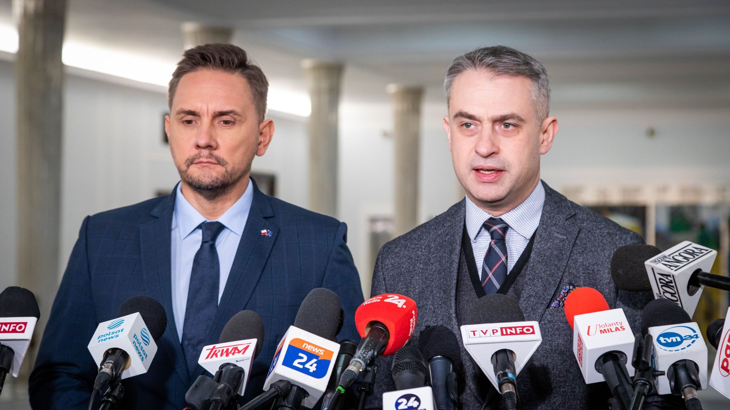 Dwóch mężczyzn w garniturach udziela konferencji prasowej, otoczonych licznymi mikrofonami z logo stacji telewizyjnych.