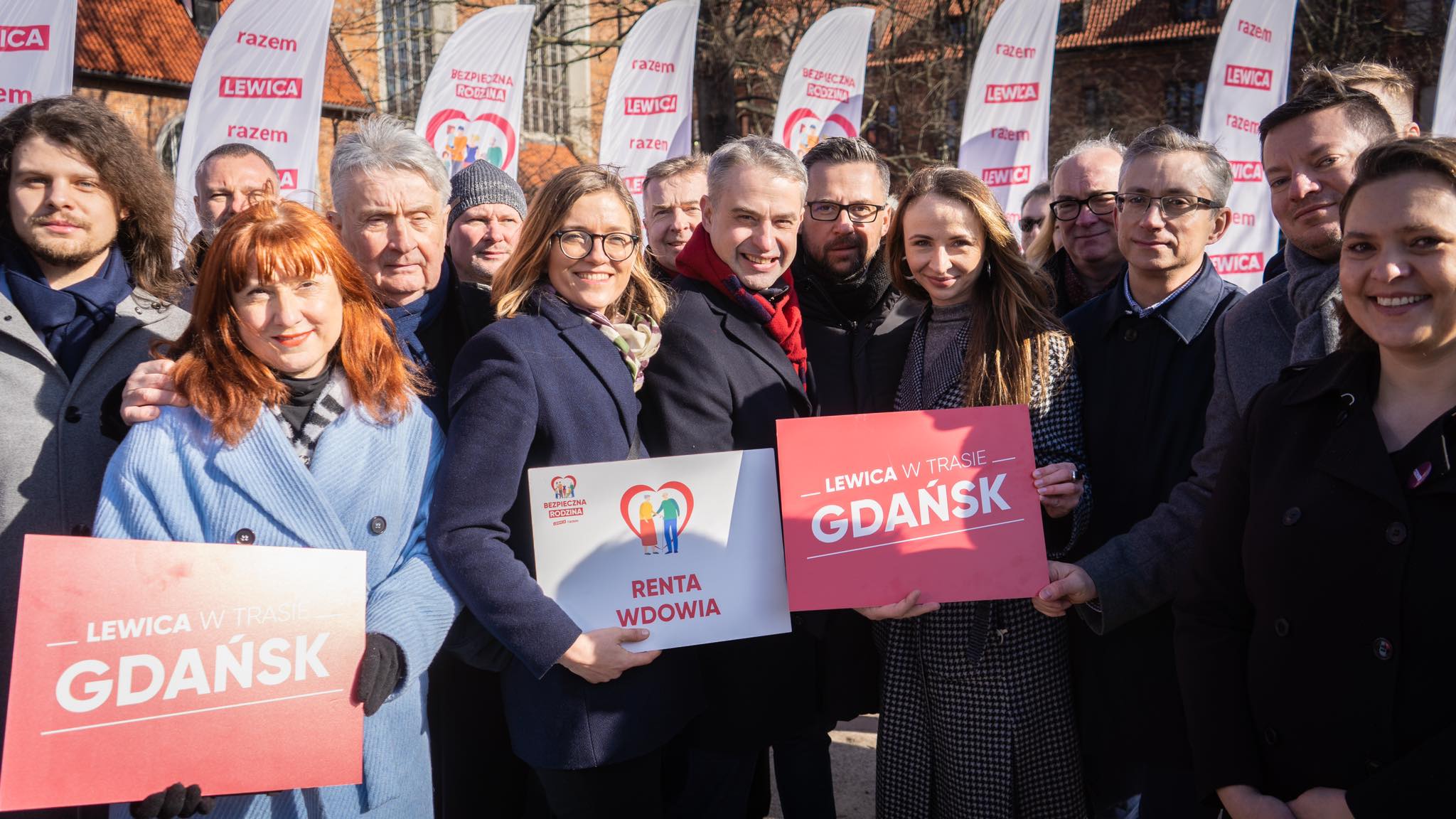 Grupa uśmiechniętych ludzi podczas wydarzenia Lewicy w Gdańsku trzyma plakaty i banery z hasłami, na tle flag.