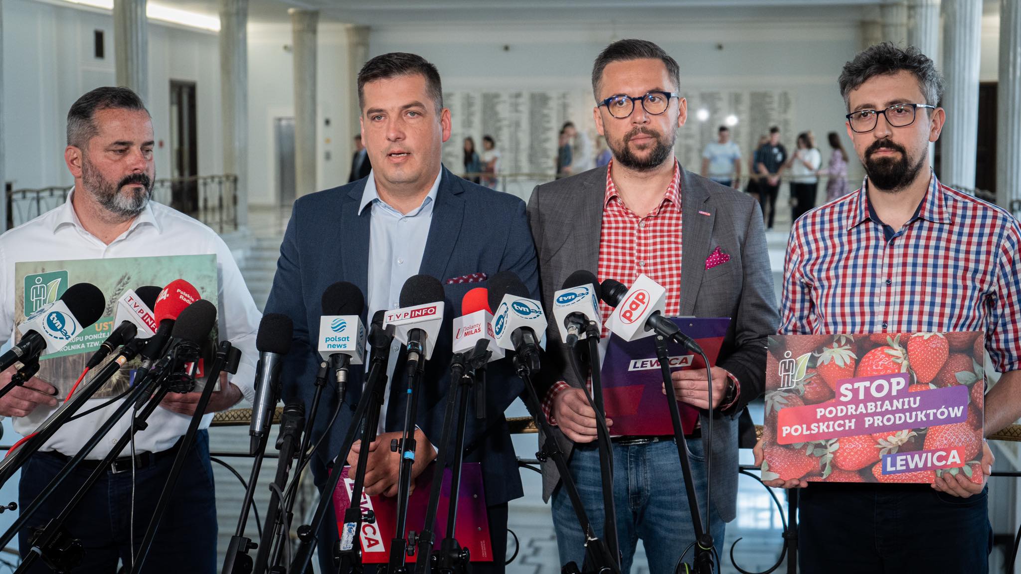 Czterech mężczyzn na konferencji prasowej stoją za licznymi mikrofonami, trzymając plakaty z hasłem STOP podrabianiu polskich produktów i logiem Lewicy.