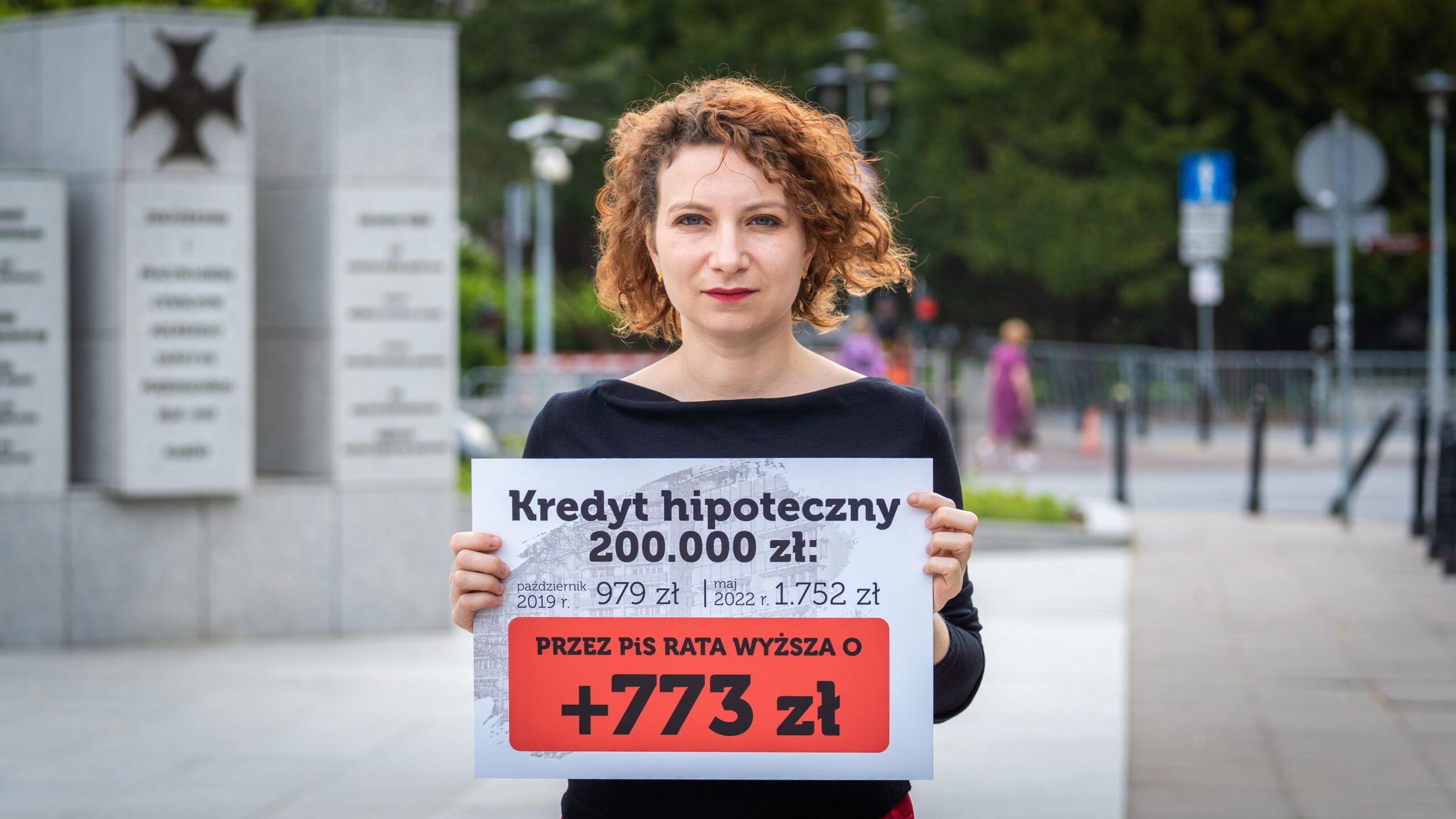 Kobieta stojąca na placu trzyma plakat z informacją o kredycie hipotecznym 200 000 zł, na tle miejskiego otoczenia z pomnikiem.