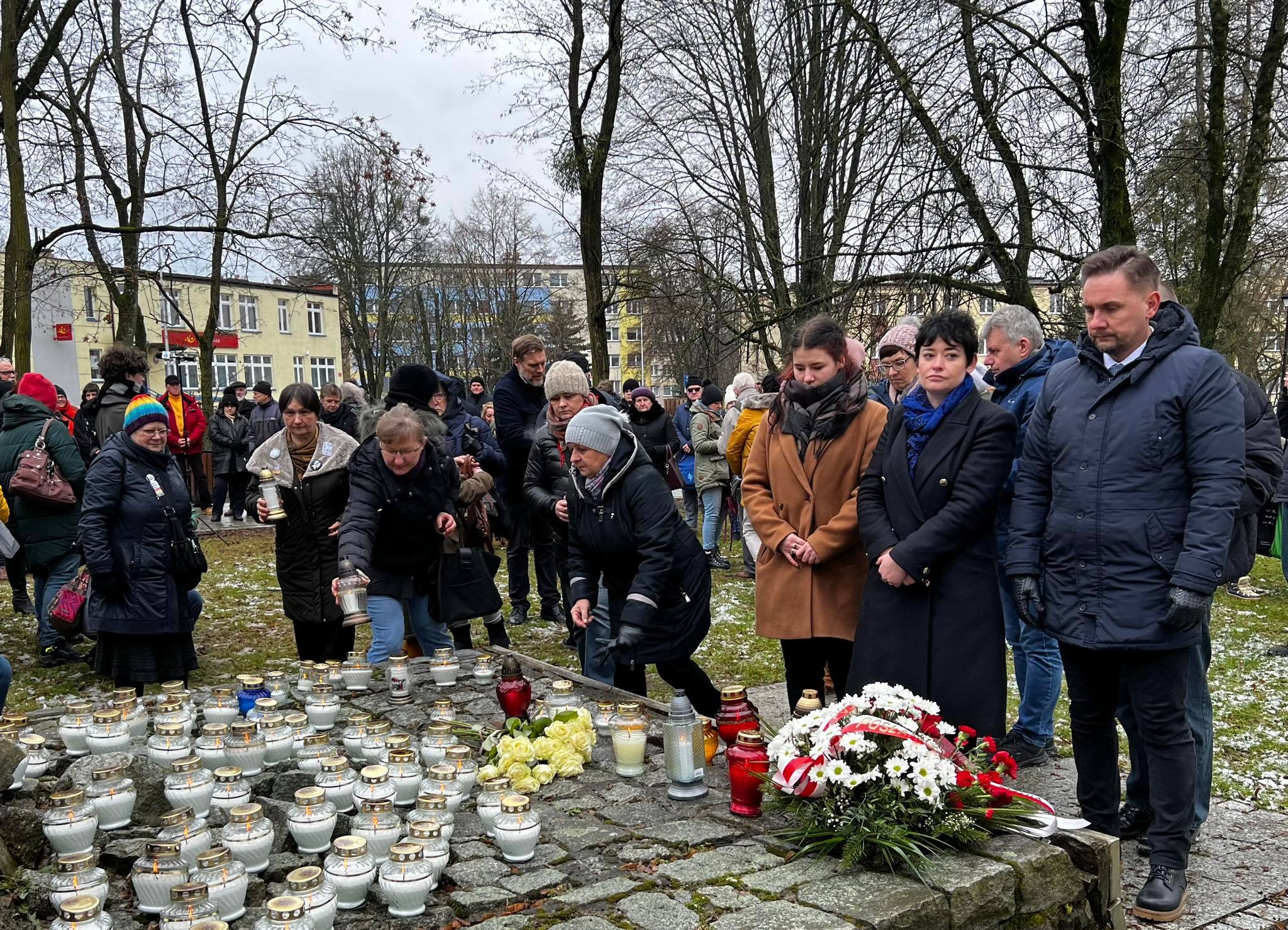 Grupa osób w zimowych kurtkach zgromadzona w Hajnowce, składa zapalone znicze i kwiaty przy miejscu pamięci upamiętniającym ofiary Romualda Rajsa „Burego”.