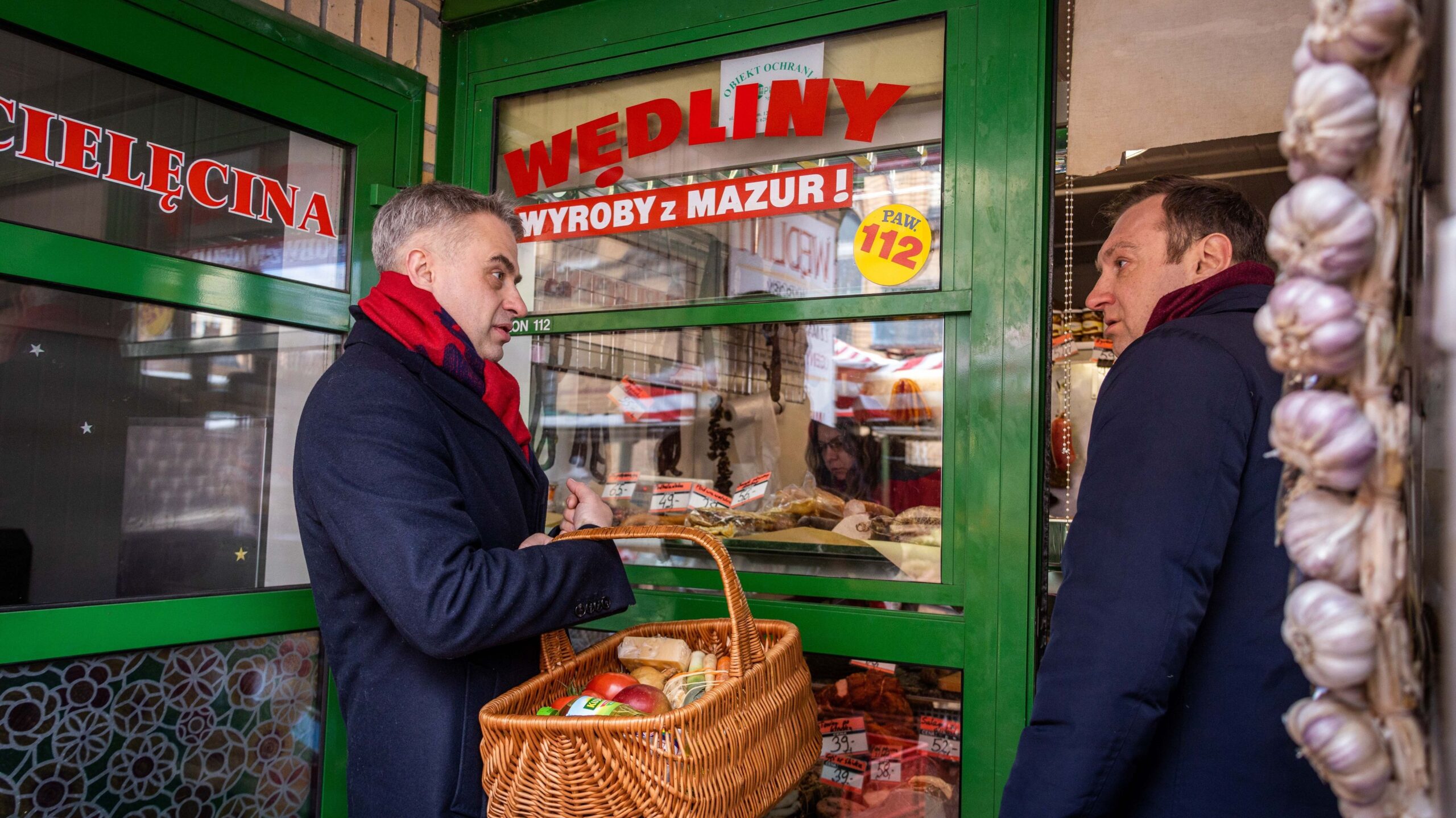 Na zdjęciu dwóch mężczyzn rozmawia przed sklepem z wędlinami, trzymając koszyk z zakupami.