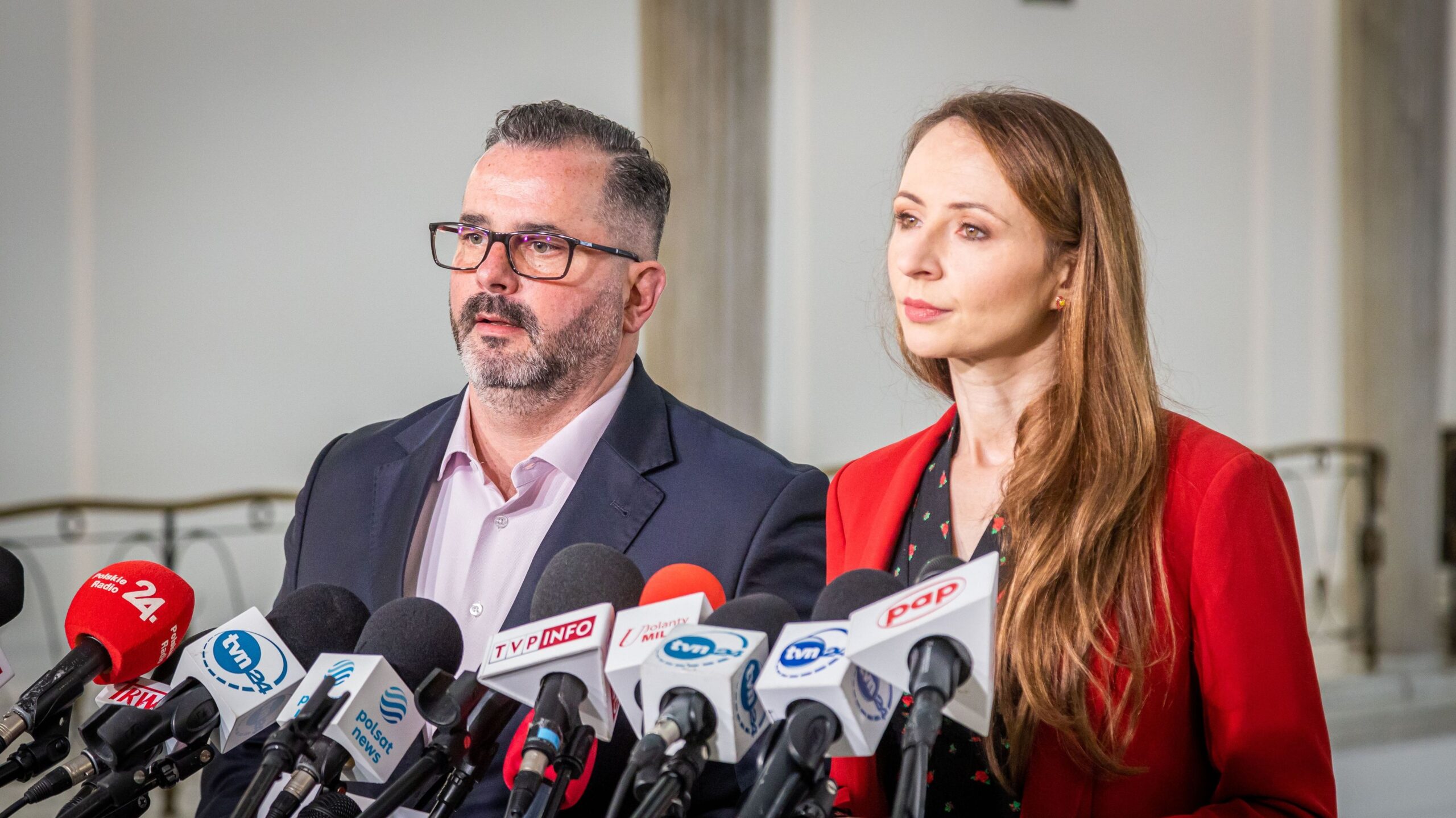 Na zdjęciu widać dwoje ludzi stojących przed mikrofonami na konferencji prasowej.