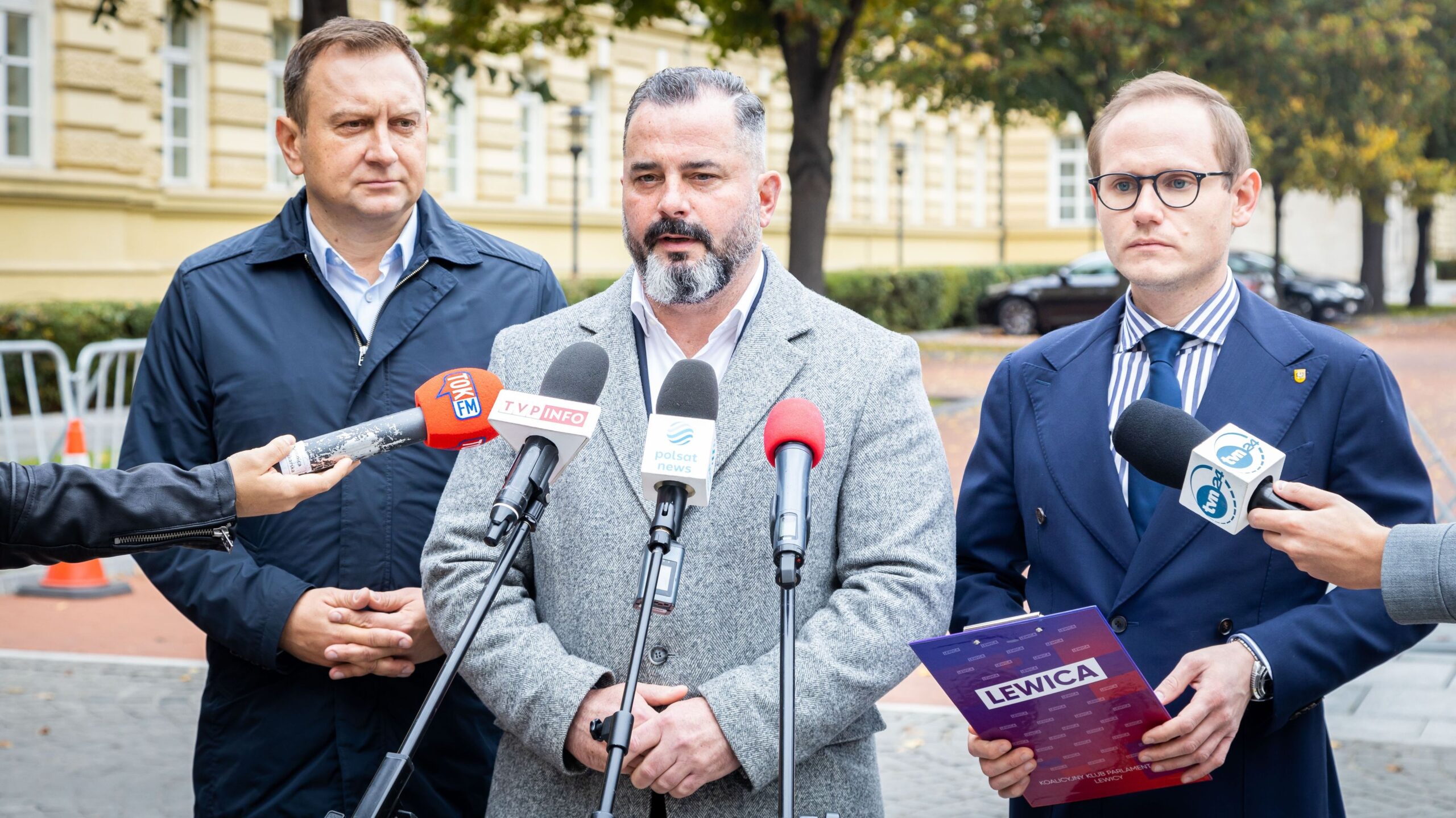 Trzej mężczyźni, w tym jeden z brodą ten w szarym płaszczu, wygłaszają oświadczenie do mediów podczas konferencji prasowej na zewnątrz.