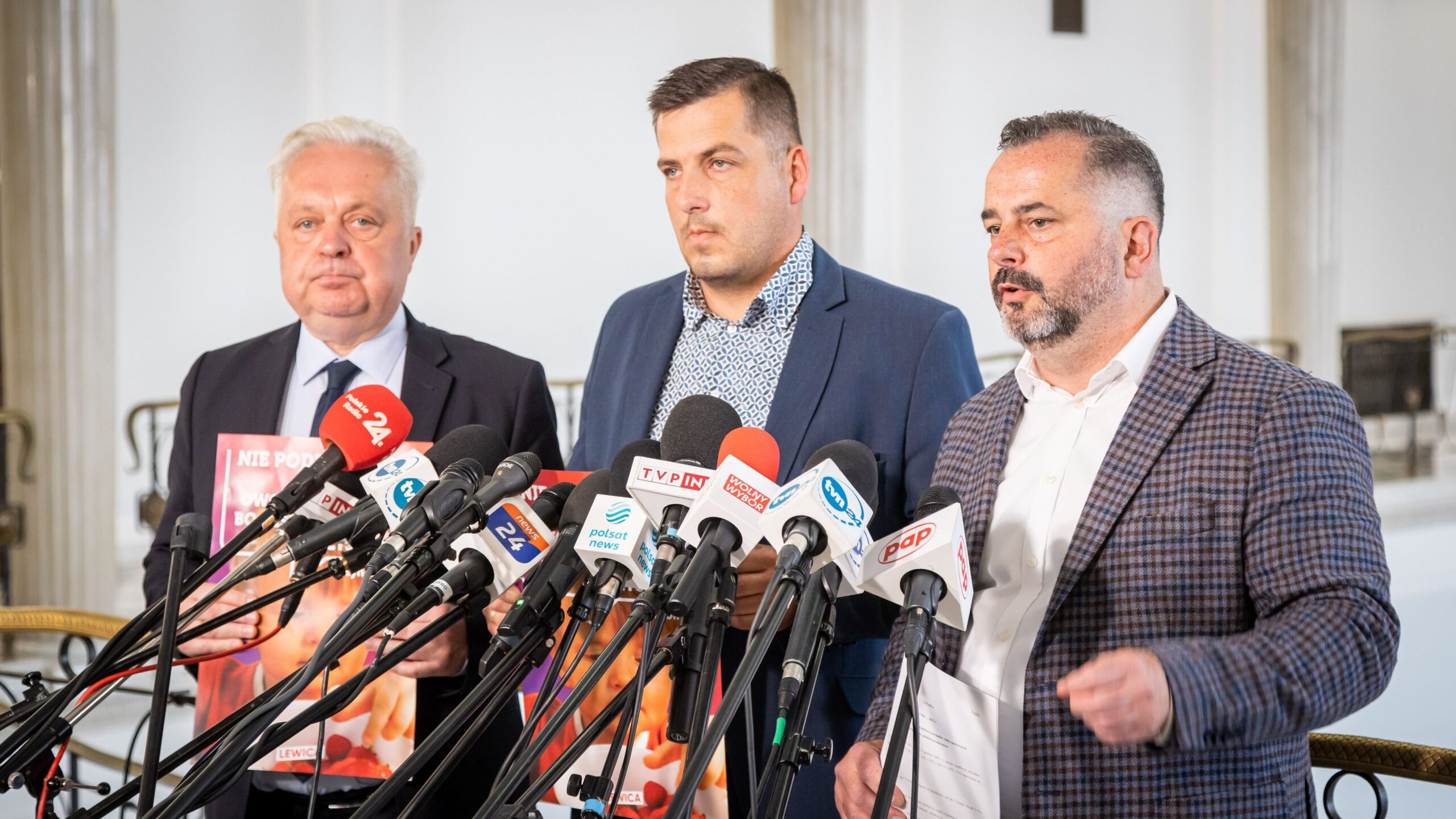 Na zdjęciu trzech mężczyzn stoi przed mikrofonami na konferencji prasowej, prezentując plakat z hasłem