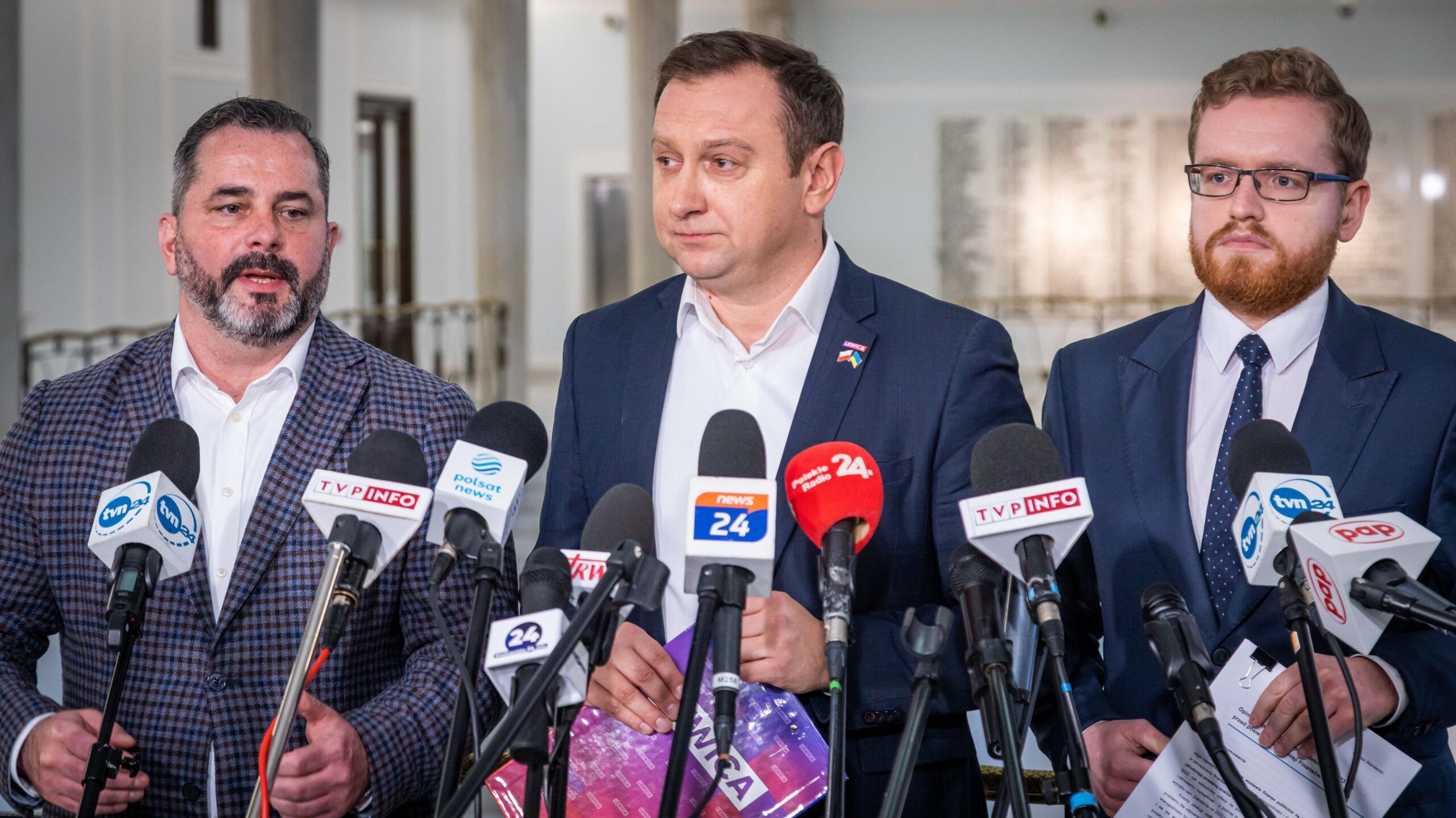 Trzej mężczyźni stoją przy mikrofonach, prowadząc konferencję prasową w dużym, jasnym pomieszczeniu.