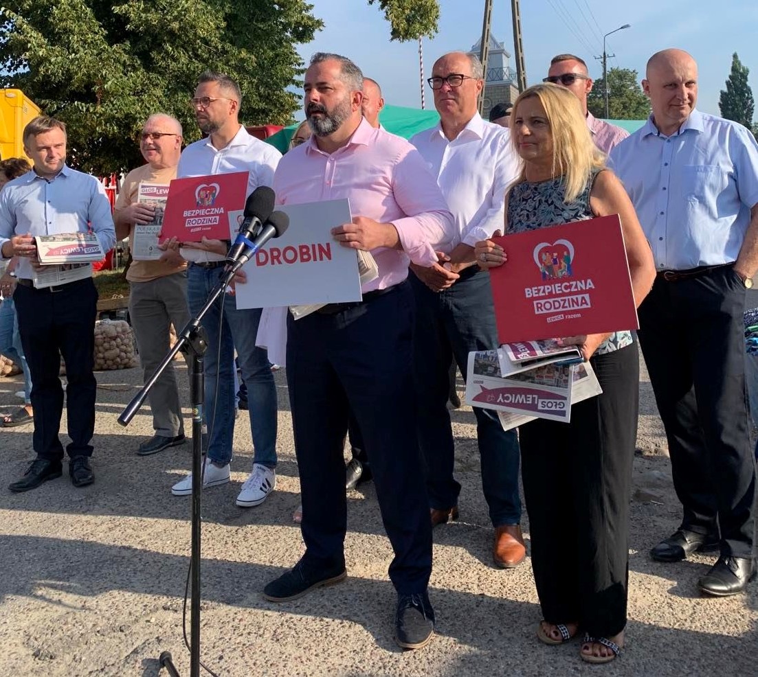 Na zdjęciu grupa ludzi stoi na zewnątrz i trzyma znaki z hasłami „DROBIN” oraz „BEZPIECZNA RODZINA” podczas wydarzenia publicznego.
