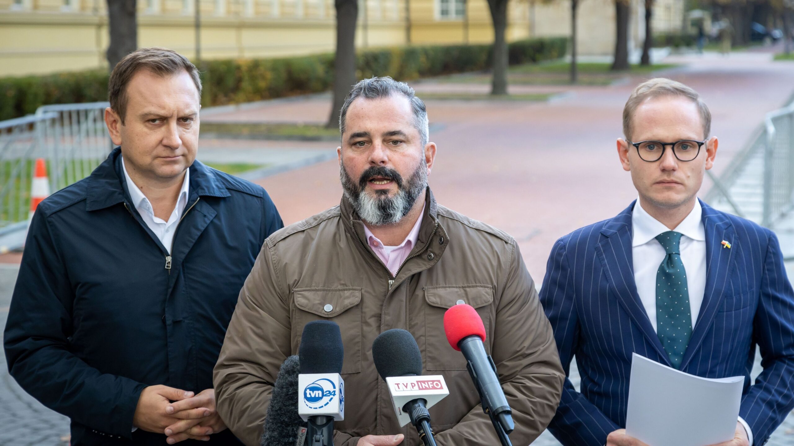 Na zdjęciu trzech mężczyzn w eleganckich ubraniach stoi w plenerze, rozmawiając do mikrofonów przed kamerami, w tle widać ulicę i drzewa.