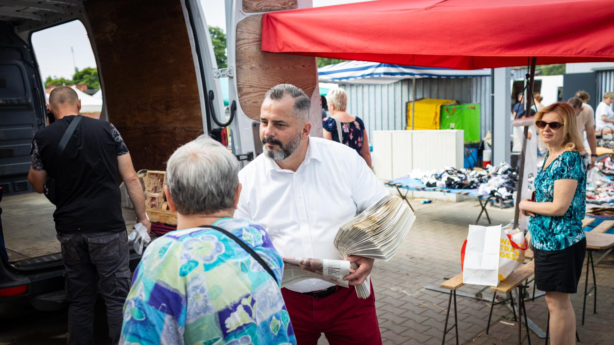 Mężczyzna w białej koszuli rozdaje gazety na targowisku, otoczony ludźmi przy stoiskach z ubraniami i stojącą po prawej kobiecą postacią w okularach przeciwsłonecznych.