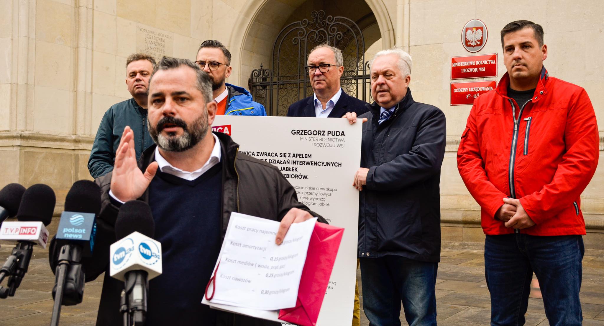 Na zdjęciu znajduje się grupa mężczyzn, w tym postać przemawiająca do mediów, w tle z tablicą z apelem o wprowadzenie działań interwencyjnych dotyczących zbiorów jabłek, przed budynkiem ministerstwa.