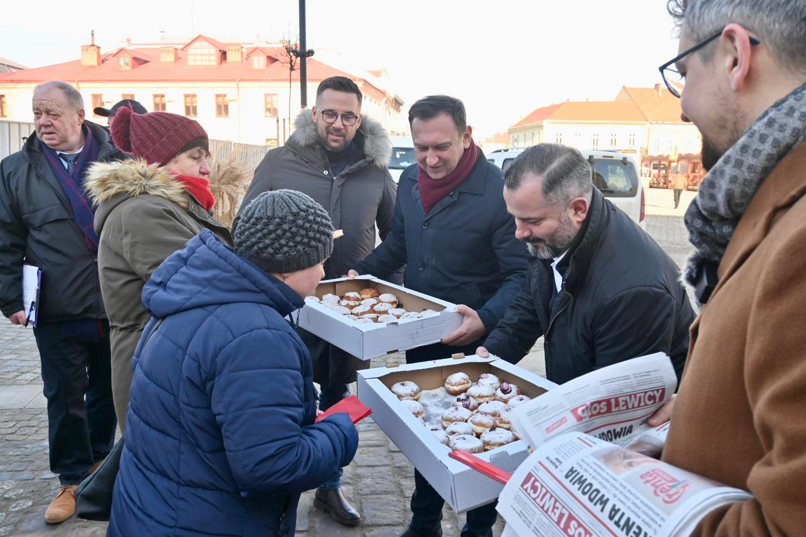 Na zdjęciu grupa osób wręcza dziecku pudełka z pączkami w zimowej scenerii.