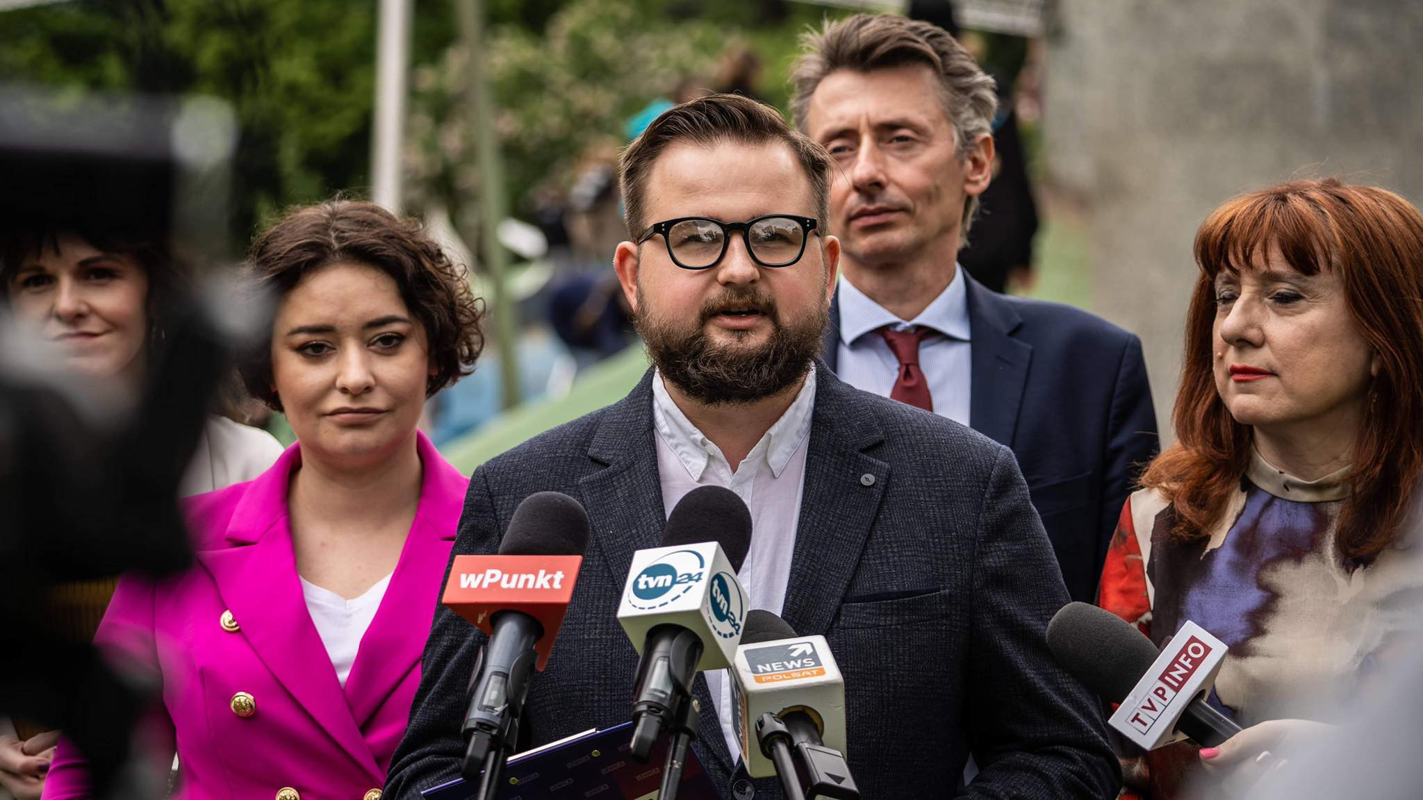 Mężczyzna w okularach i ciemnym garniturze przemawia na konferencji prasowej związanej z Pakietem Zdrowego Dziecka, otoczony reporterami i mikrofonami z logotypami mediów.