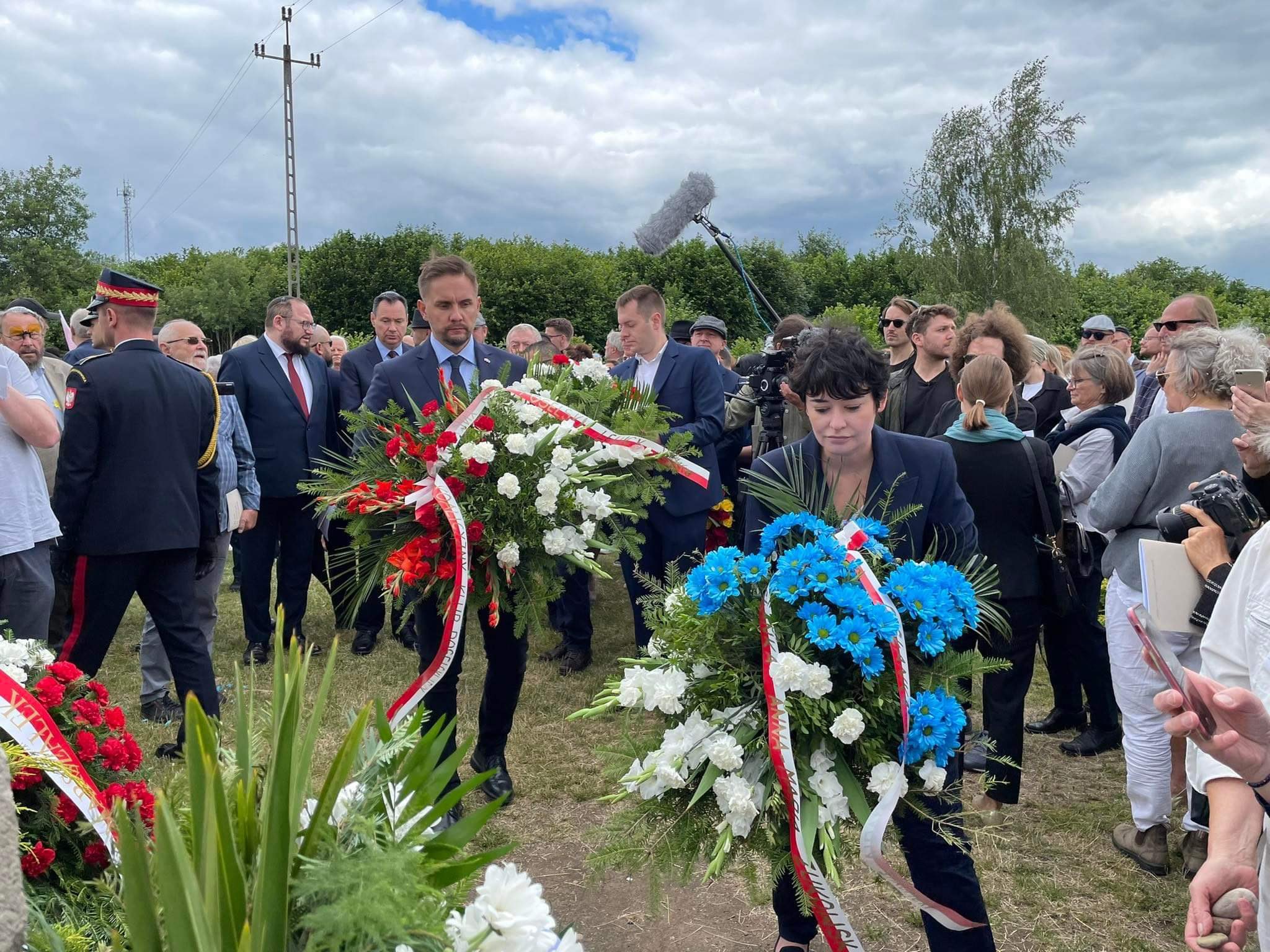 Zdjęcie przedstawia grupę ludzi uczestniczących w uroczystości złożenia kwiatów, wśród których znajdują się osoby w garniturach niosące wieńce, a w tle widoczne są zebrane tłumy i chmurne niebo.