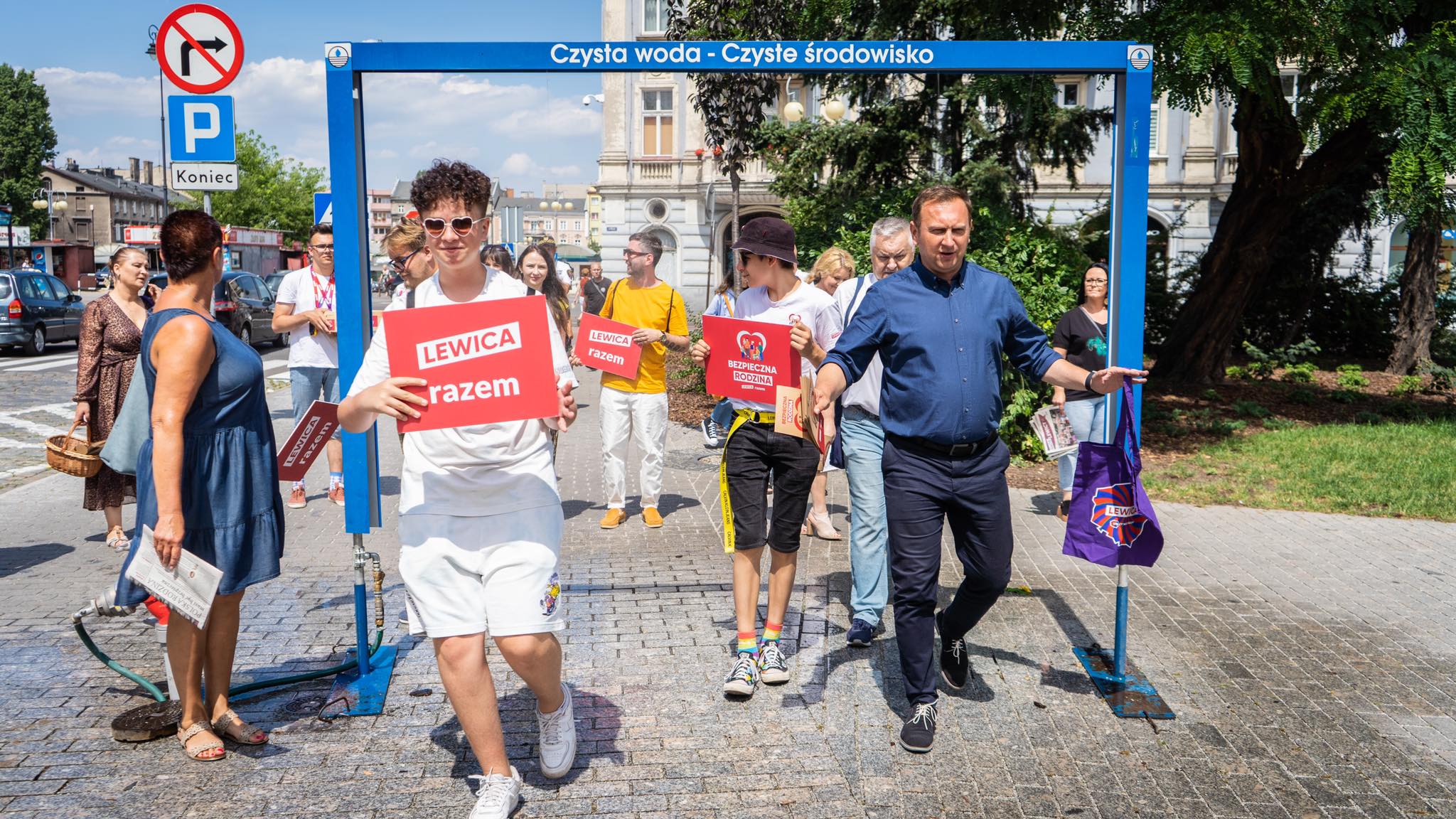 Grupa uczestników demonstracji politycznej przechodzi pod niebieską bramą z napisem „Czysta woda - Czyste środowisko”, trzymając czerwone plakaty z hasłem „Lewica razem”.