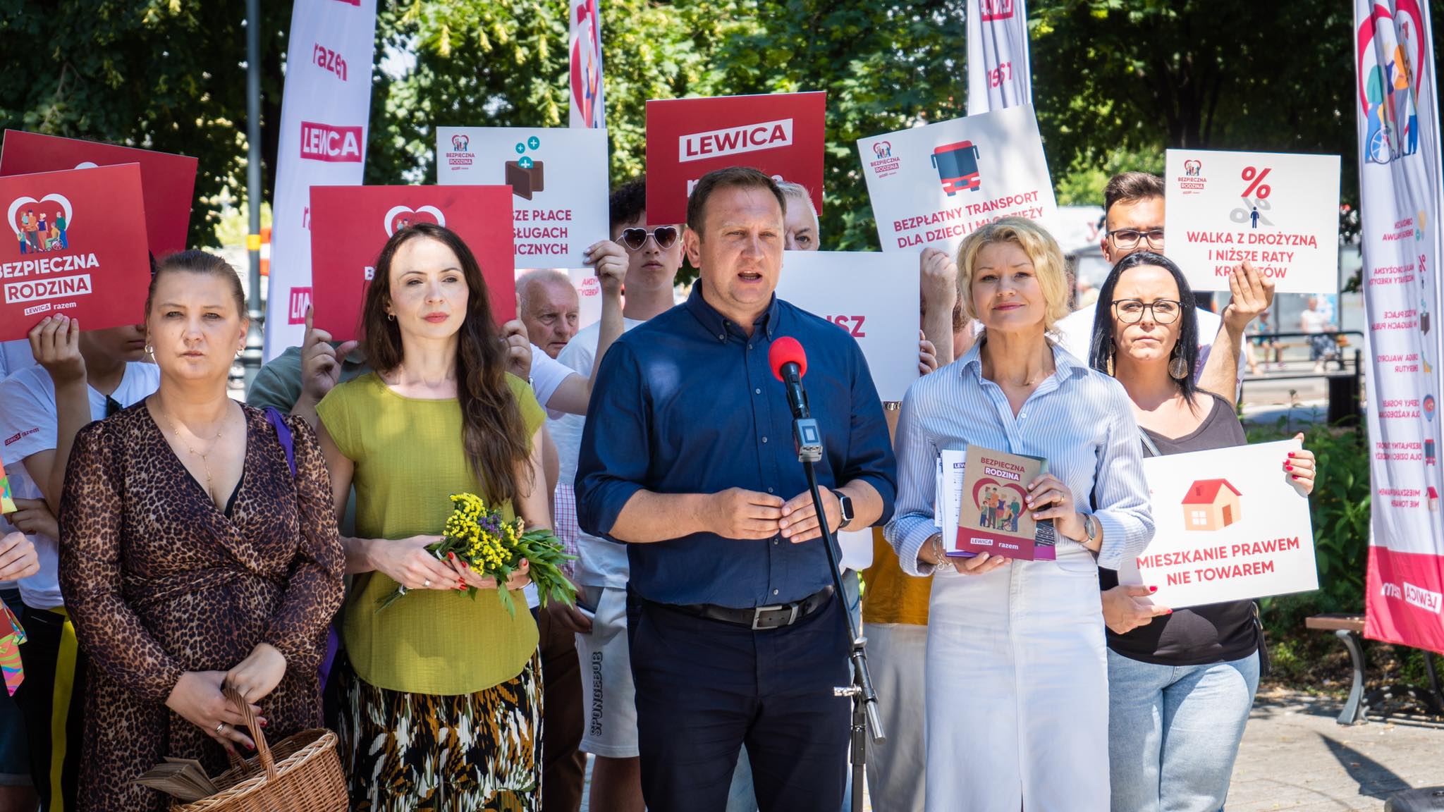 Grupa uczestników wiecu Lewicy trzyma plakaty i banery z hasłami, a mężczyzna w środku przemawia do mikrofonu.