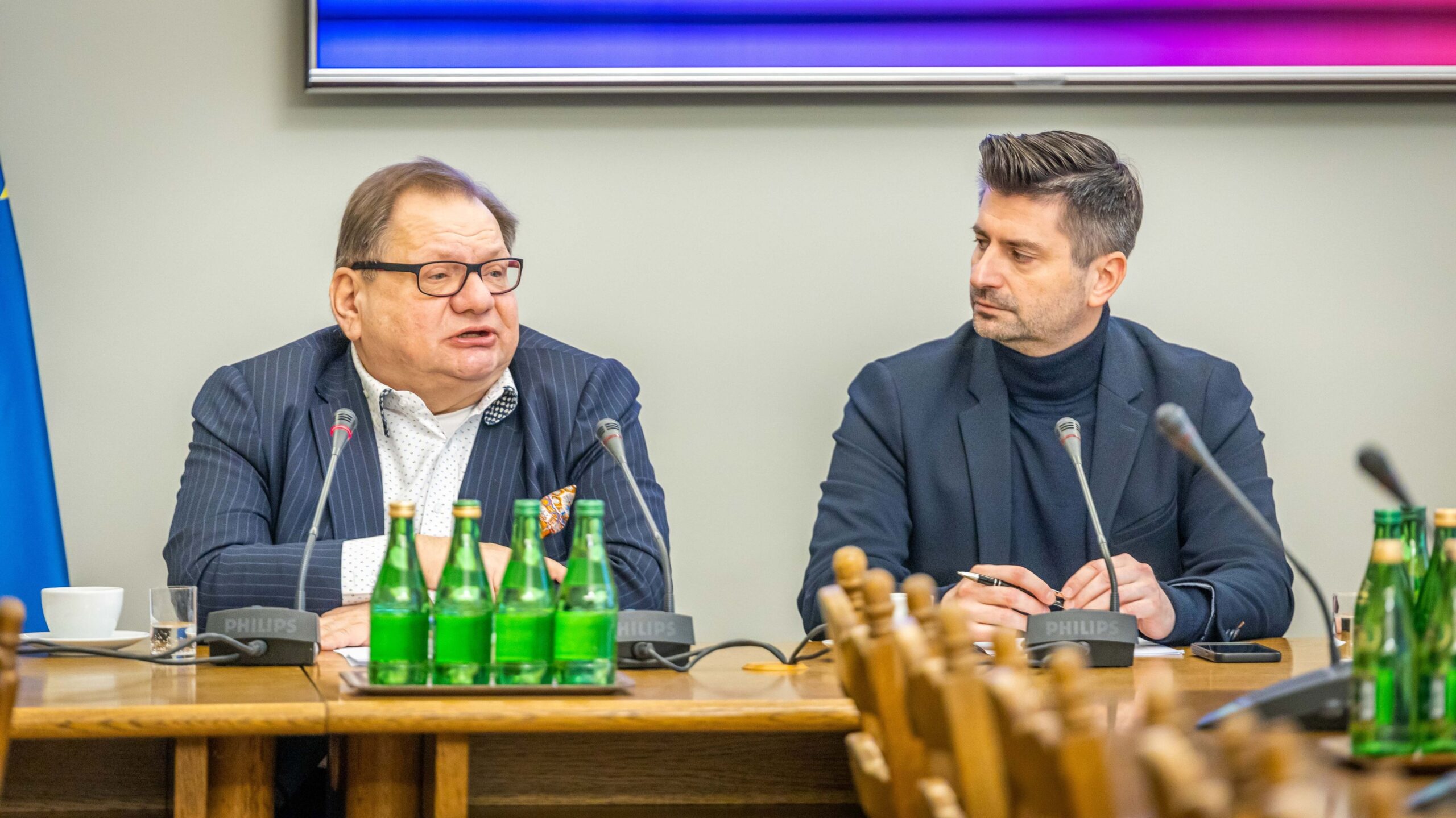 Dwóch mężczyzn w garniturach siedzi przy długim stole podczas konferencji prasowej, z mikrofonami i zielonymi butelkami wody na blacie.