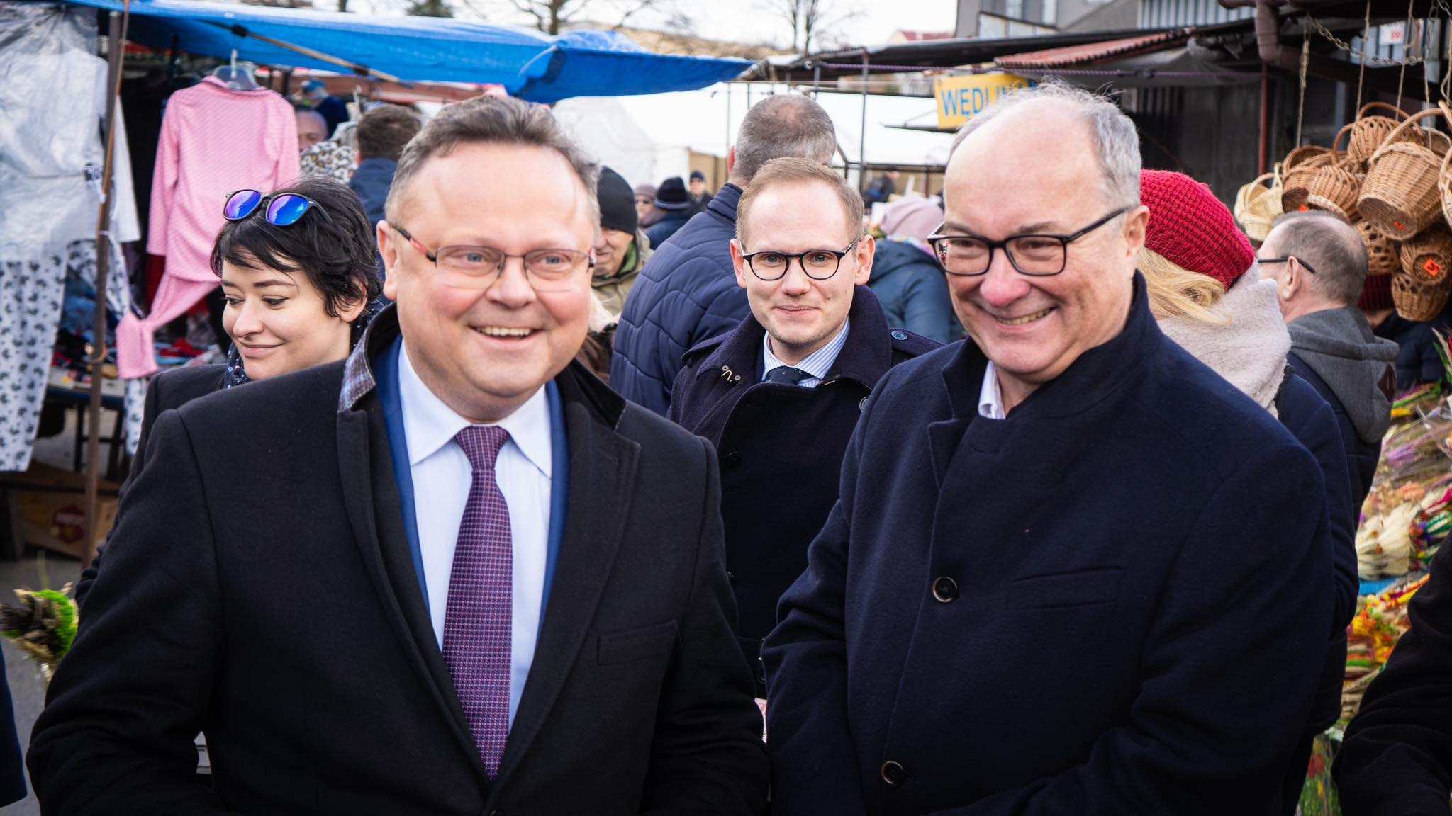 Na zdjęciu widać grupę mężczyzn uśmiechających się i rozmawiających na rynku, w otoczeniu straganów z różnymi towarami.