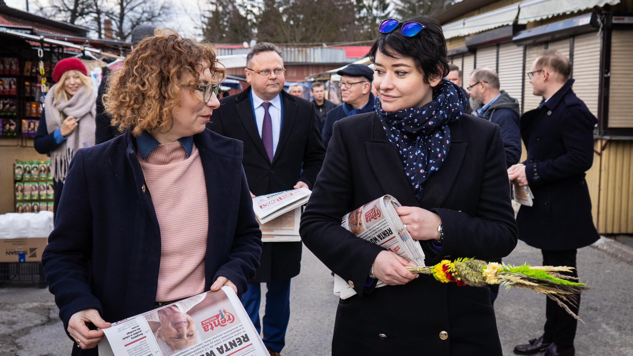 Na zdjęciu dwie kobiety rozmawiają na targu, trzymając gazety, podczas gdy w tle widać innych ludzi i stoiska handlowe.
