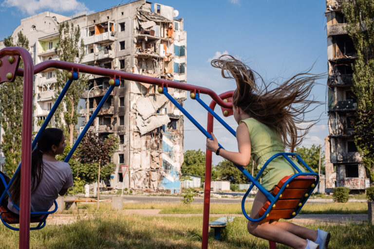 Dwie dziewczynki bawią się na huśtawce w parku, a w tle widać zniszczony blok mieszkalny.