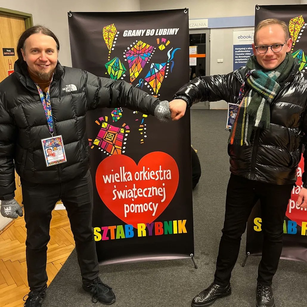 Dwóch mężczyzn w czarnych kurtkach puchowych wykonuje gest stuknięcia pięściami przed bannerem Wielkiej Orkiestry Świątecznej Pomocy.