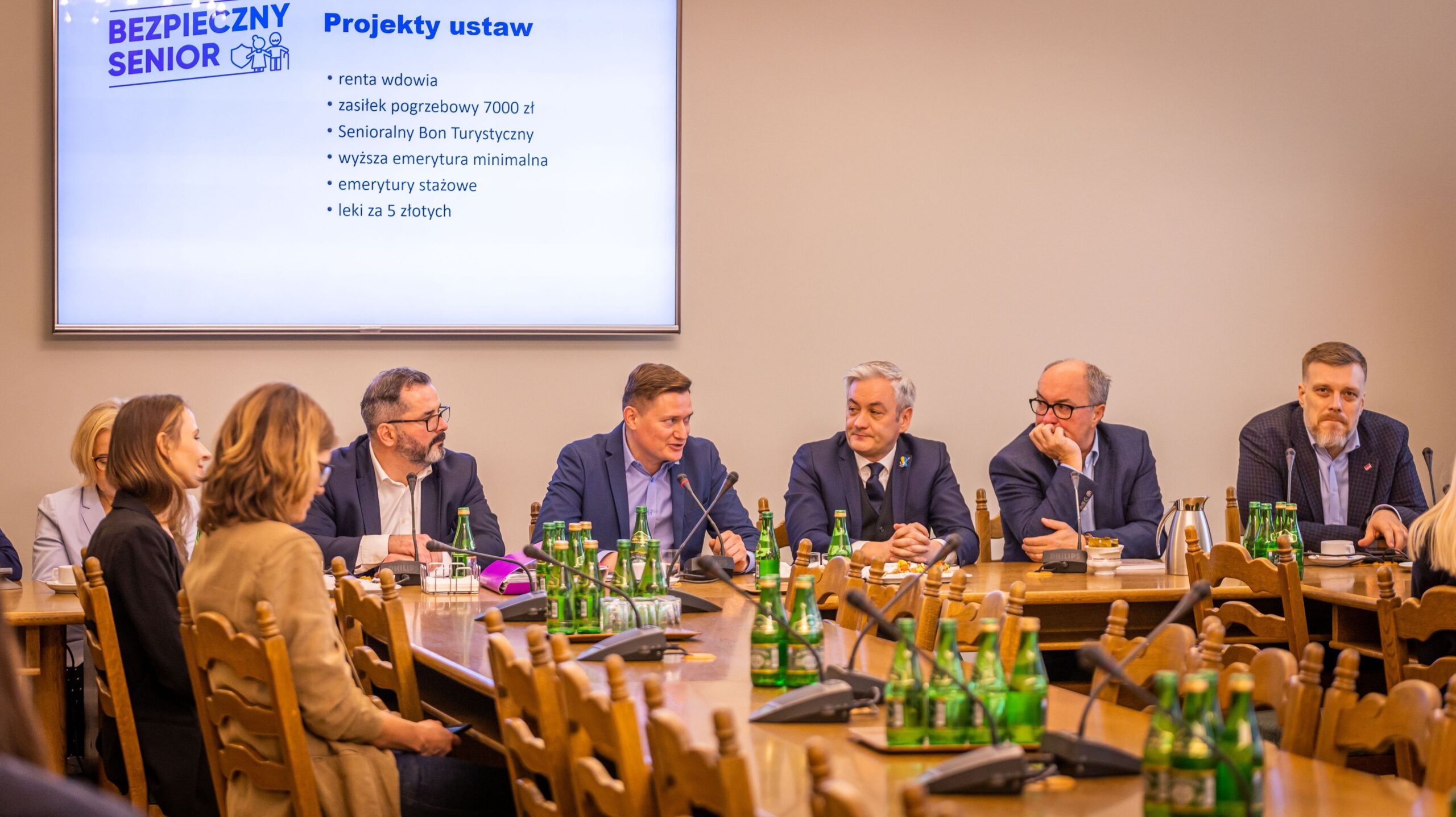 Na zdjęciu widać spotkanie członków komitetu, podczas którego omawiane są projekty ustaw dotyczące wsparcia dla seniorów, z prezentacją na ekranie w tle.