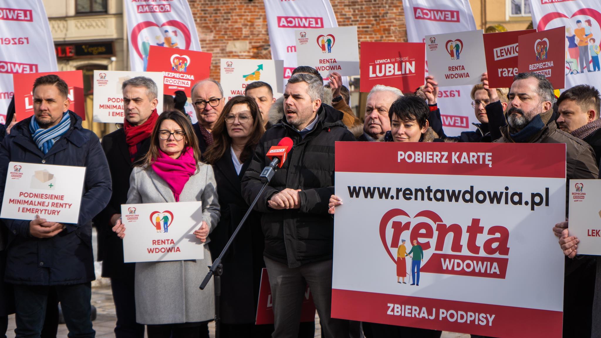 Grupa osób na demonstracji na Lubelszczyźnie trzyma plakaty i banery dotyczące renty wdowiej i bezpiecznej rodziny, podczas gdy mężczyzna w czarnym płaszczu przemawia do mikrofonu.