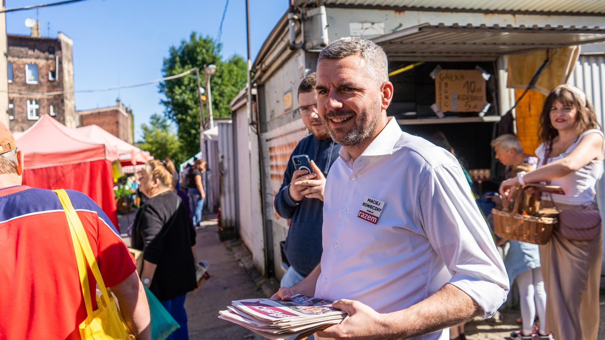Uśmiechnięty mężczyzna w białej koszuli rozdaje gazety na targu plenerowym, w tle stoją stragany i przechodnie.