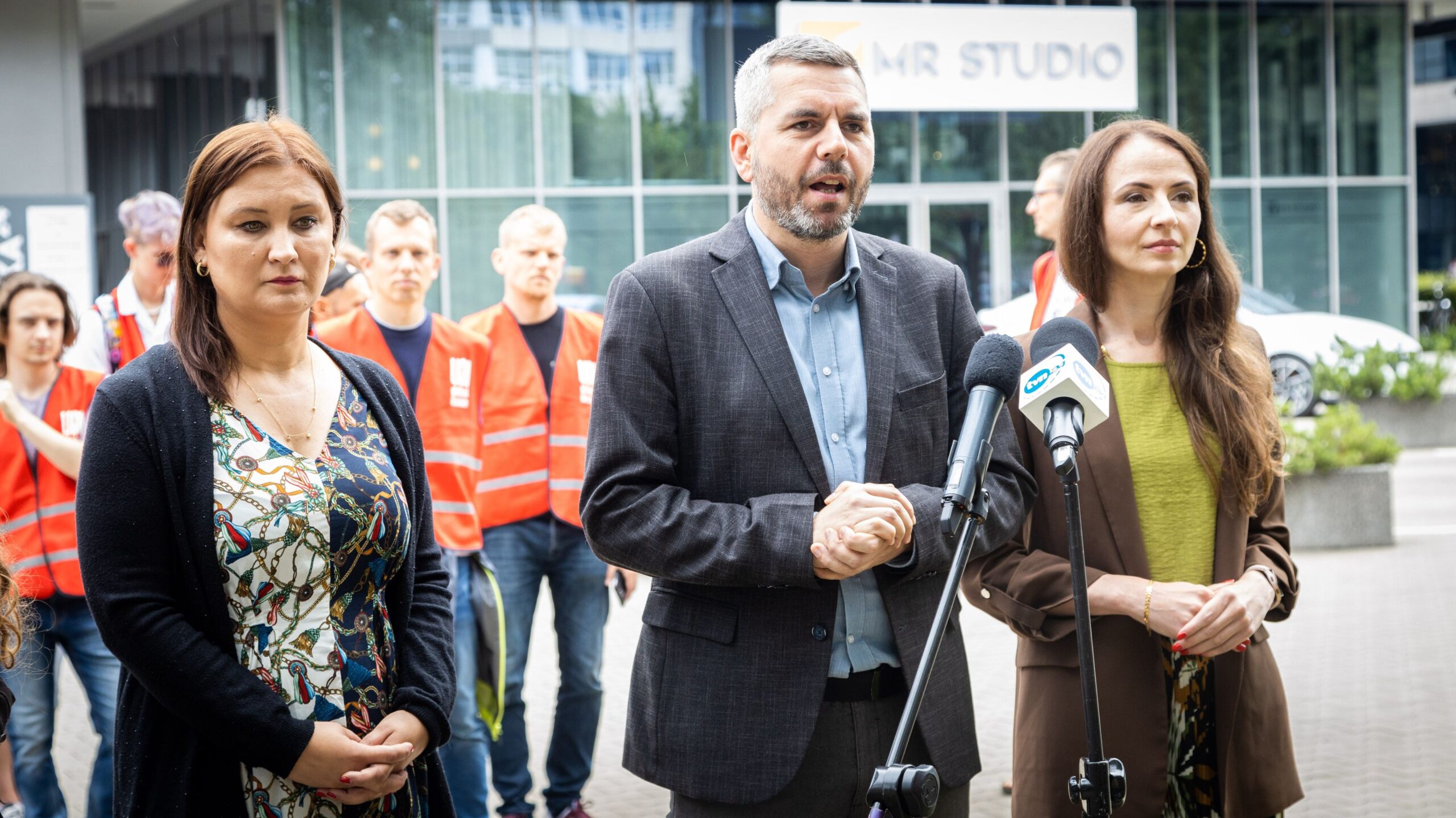 Na zdjęciu widać trzech mówiących do mediów ludzi, w tym dwóch w garniturach, stojących przed grupą osób ubranych w pomarańczowe kamizelki.