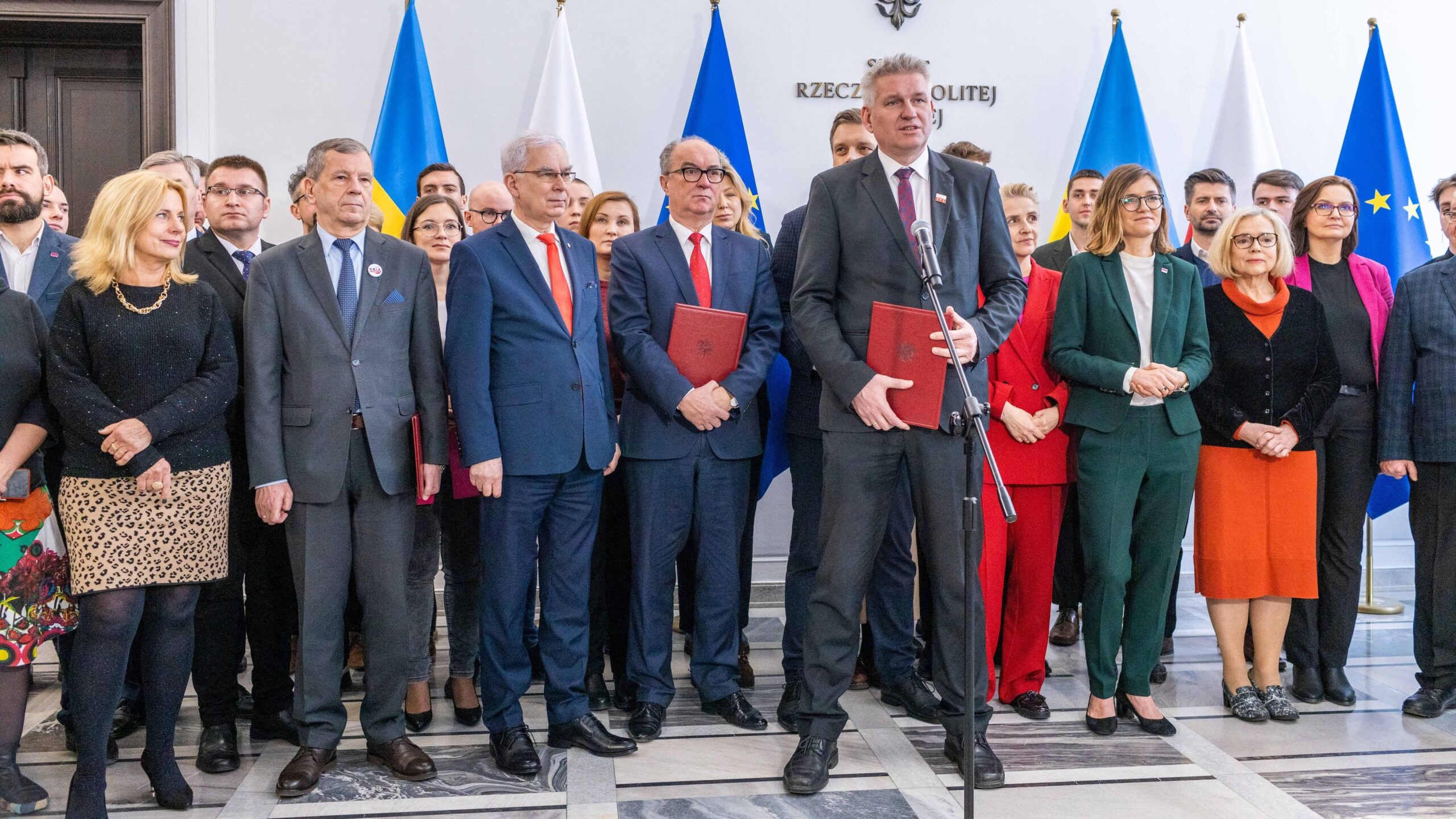 Na zdjęciu widoczna jest grupa ludzi w formalnych strojach podczas wydarzenia w towarzystwie flag i dokumentów, z jednym mężczyzną przemawiającym do mikrofonu.