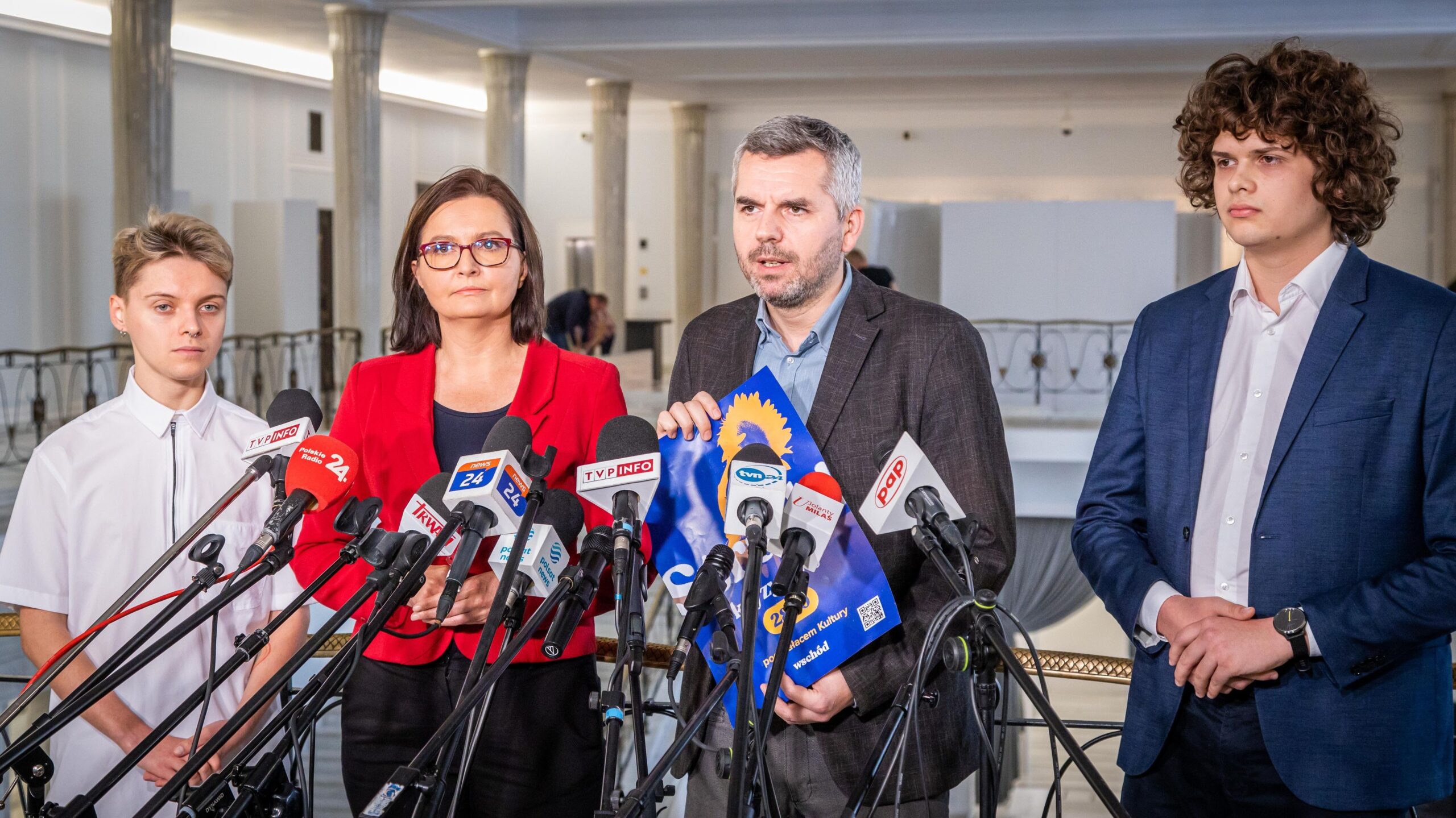 Na zdjęciu widać czterech ludzi stojących przed mikrofonami na konferencji prasowej, z jednym z nich trzymającym dokument, w tle eleganckie wnętrze.