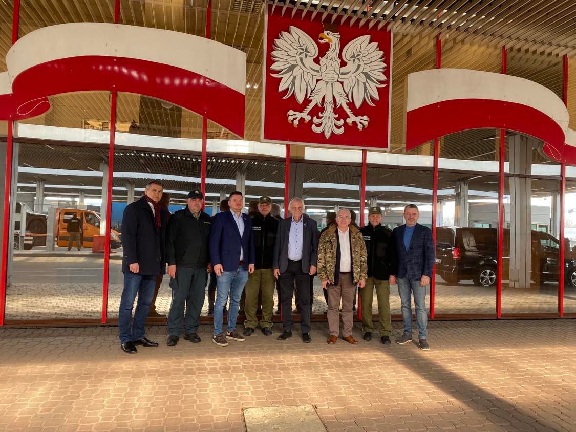 Grupa mężczyzn stojących przed budynkiem ozdobionym dużym białym orłem na czerwonym tle i polskimi dekoracjami, podczas wizyty delegacji parlamentarnej w Łucku.