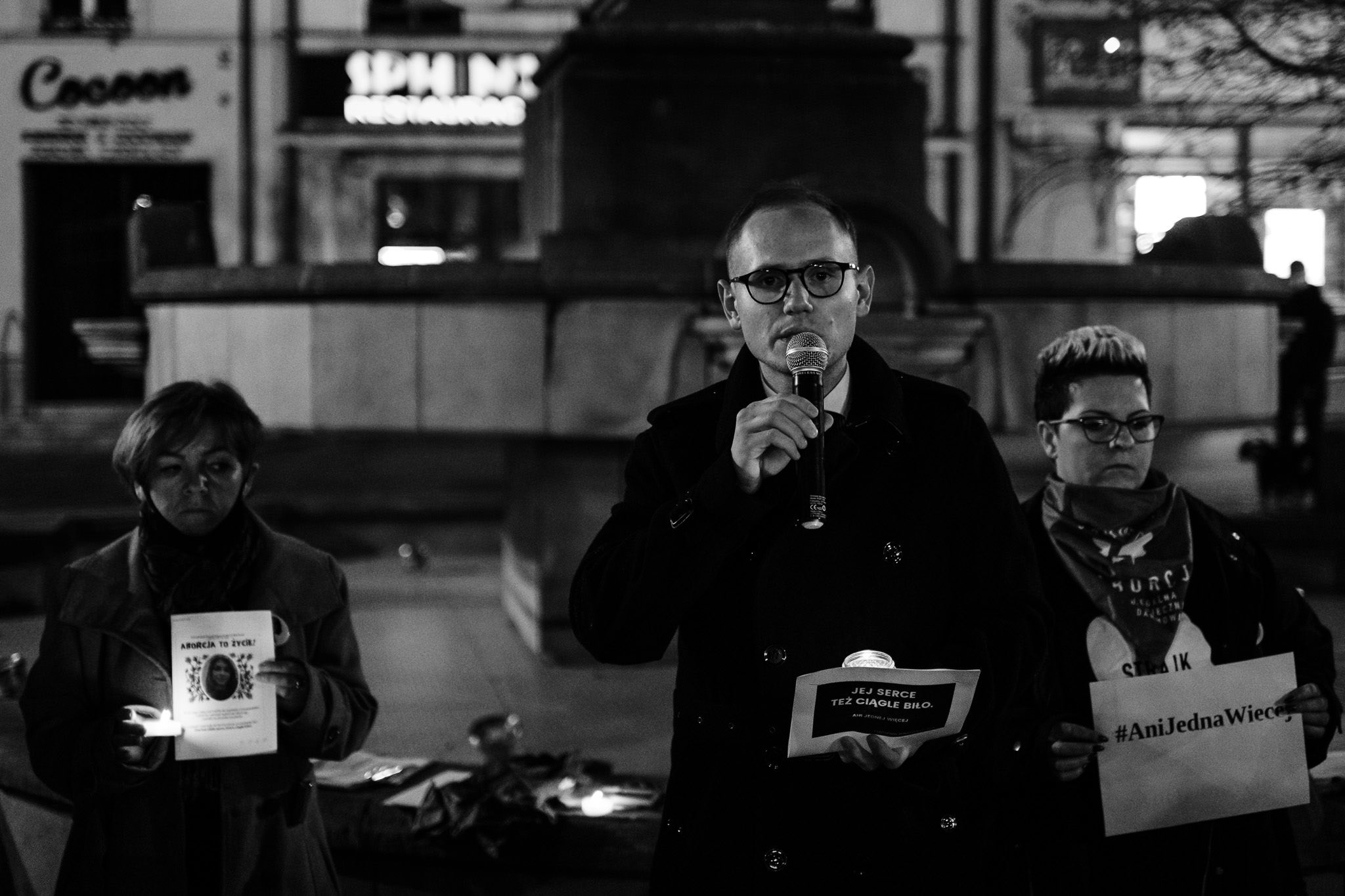 Czarno-białe zdjęcie z wiecu na placu przed pomnikiem: pośrodku mężczyzna w okularach przemawia do mikrofonu, po jego lewej stoi kobieta trzymająca świecę i ulotkę, a po prawej kolejna kobieta z plakatem z hasłem „Ani Jednej Więcej”.