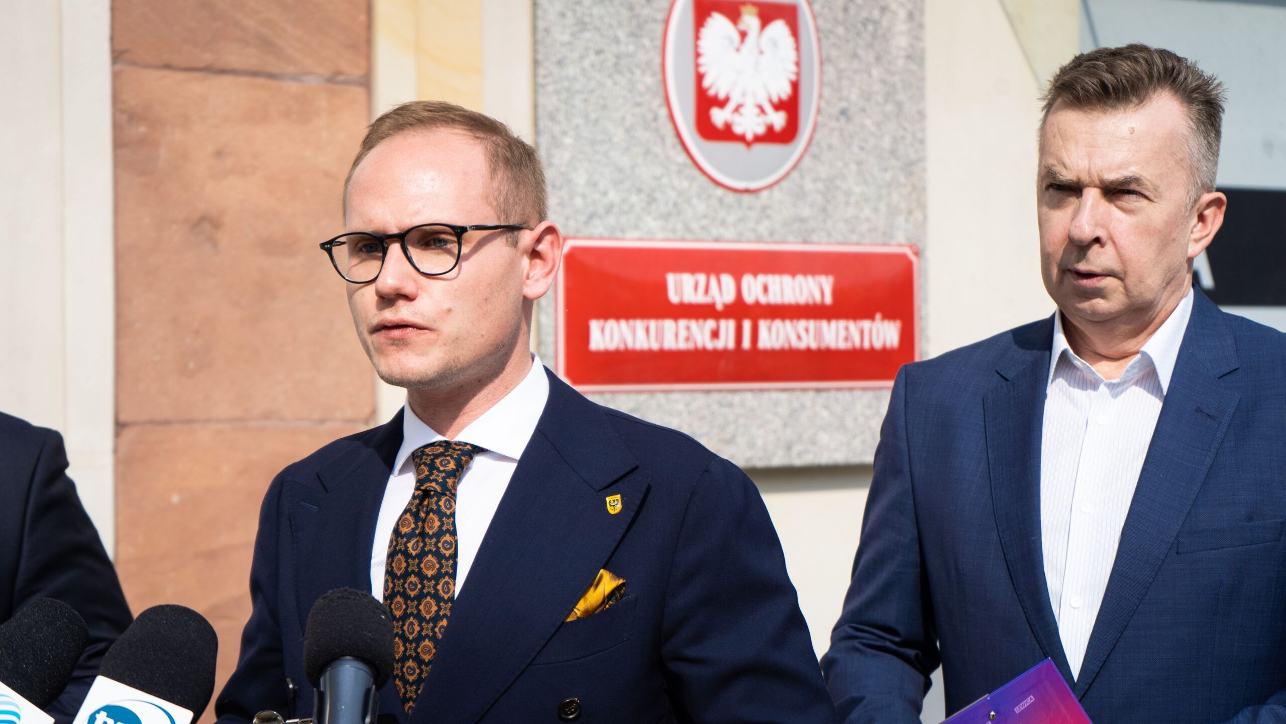 Na zdjęciu znajduje się dwóch mężczyzn w garniturach stojących przed siedzibą Urzędu Ochrony Konkurencji i Konsumentów, jeden z nich wygłasza oświadczenie do mikrofonów.