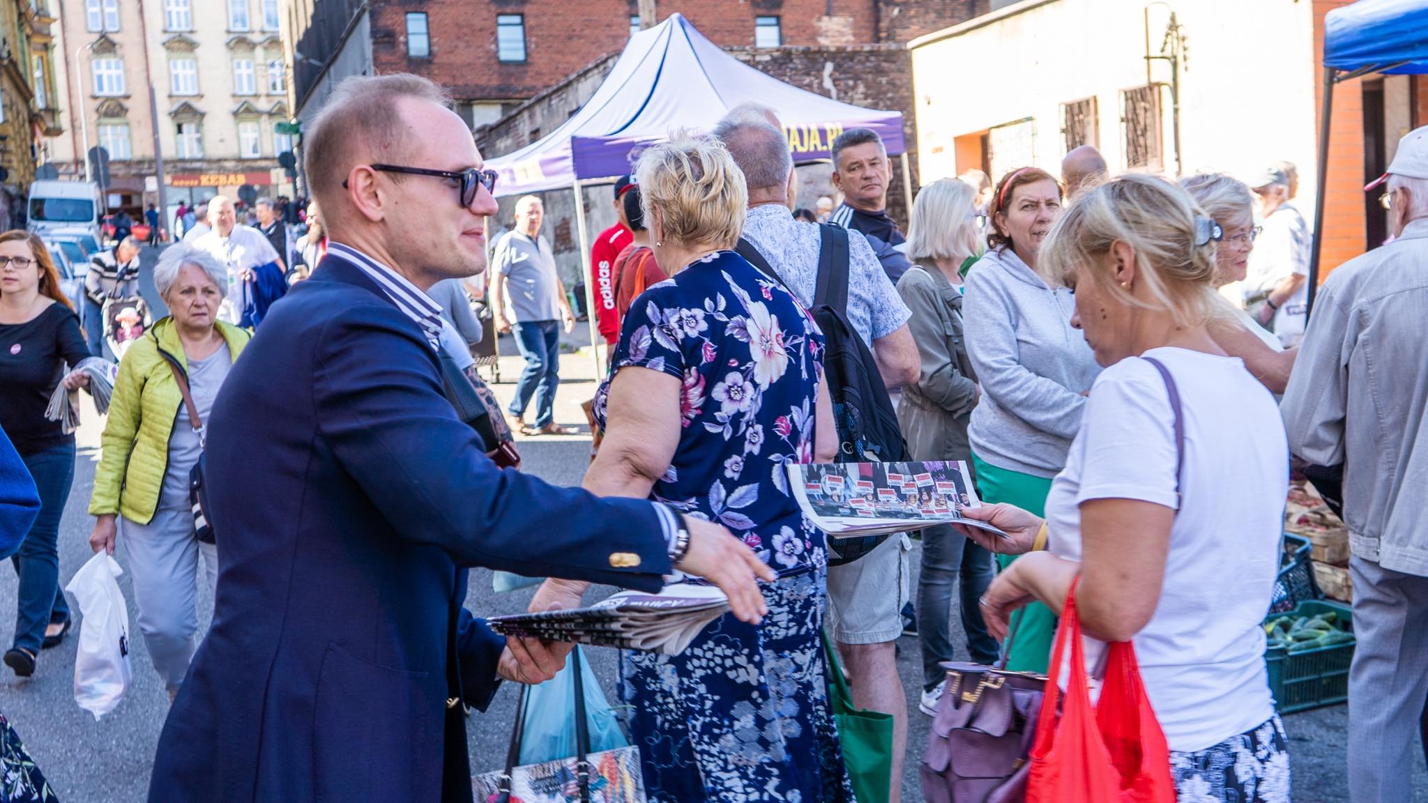 Mężczyzna w granatowym garniturze rozdaje ulotki na zatłoczonej ulicy podczas wydarzenia „Bezpieczna Rodzina”, wśród tłumu ludzi przy stoiskach i namiotach.