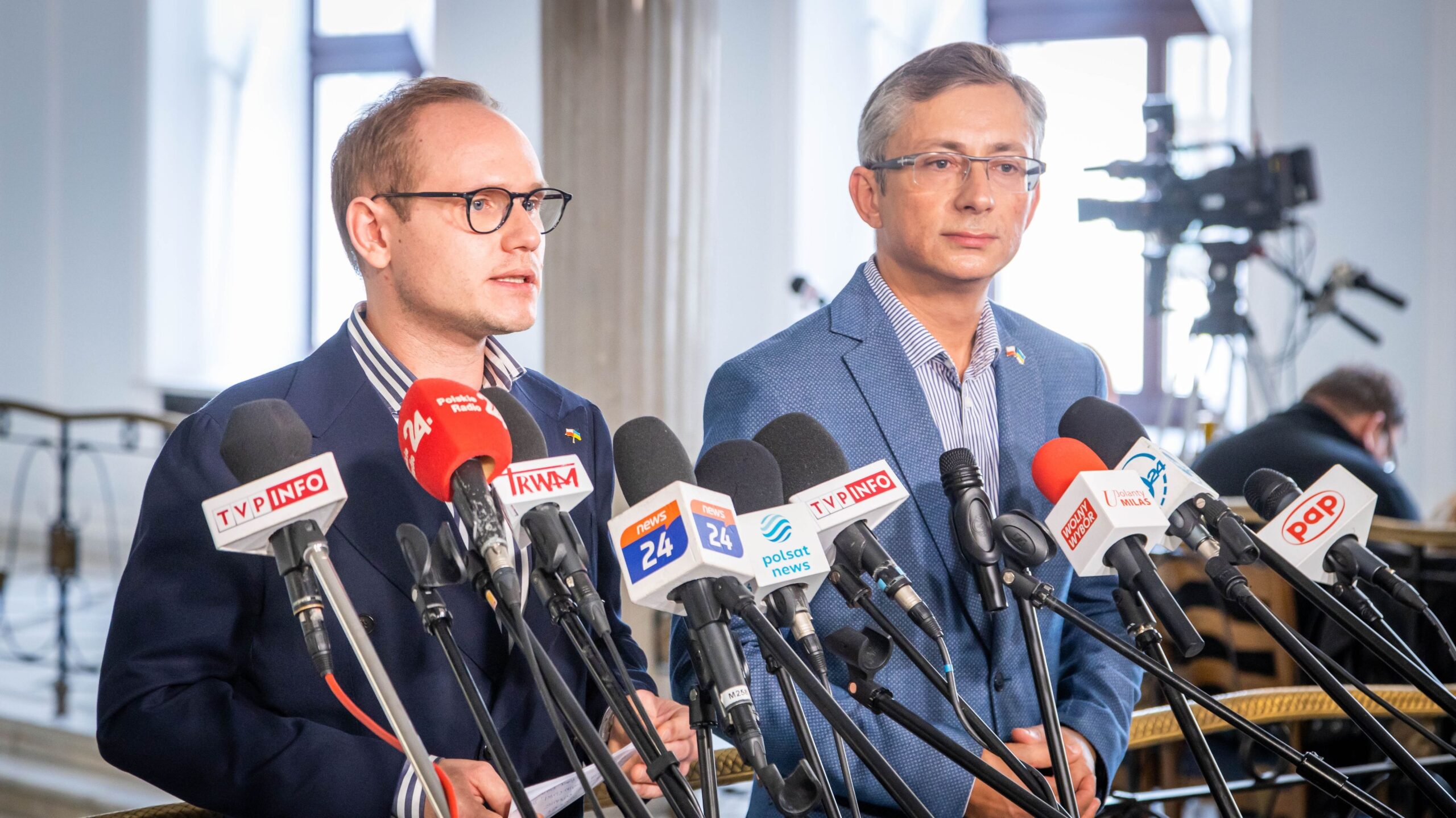 Dwaj mężczyźni stoją przy mikrofonach i prowadzą konferencję prasową w jasnym wnętrzu.