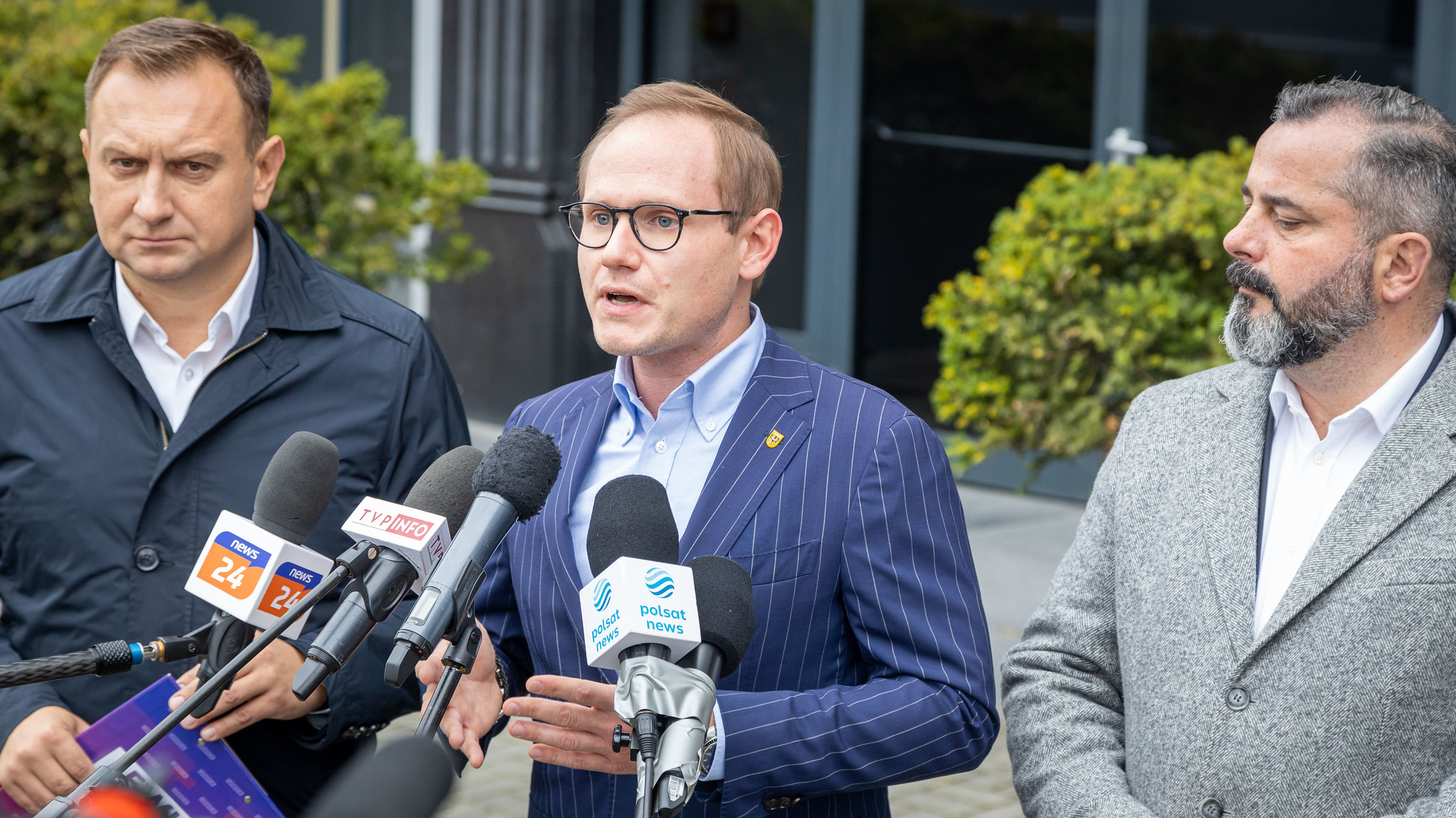 Na zdjęciu trzech mężczyzn stoi przed mikrofonami podczas konferencji prasowej, omawiając ważne kwestie.