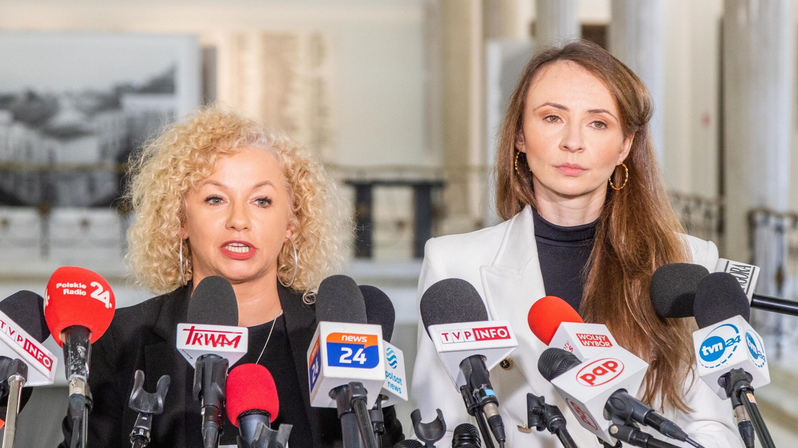 Dwie kobiety stoją przy mikrofonach podczas konferencji prasowej w przestronnej sali.