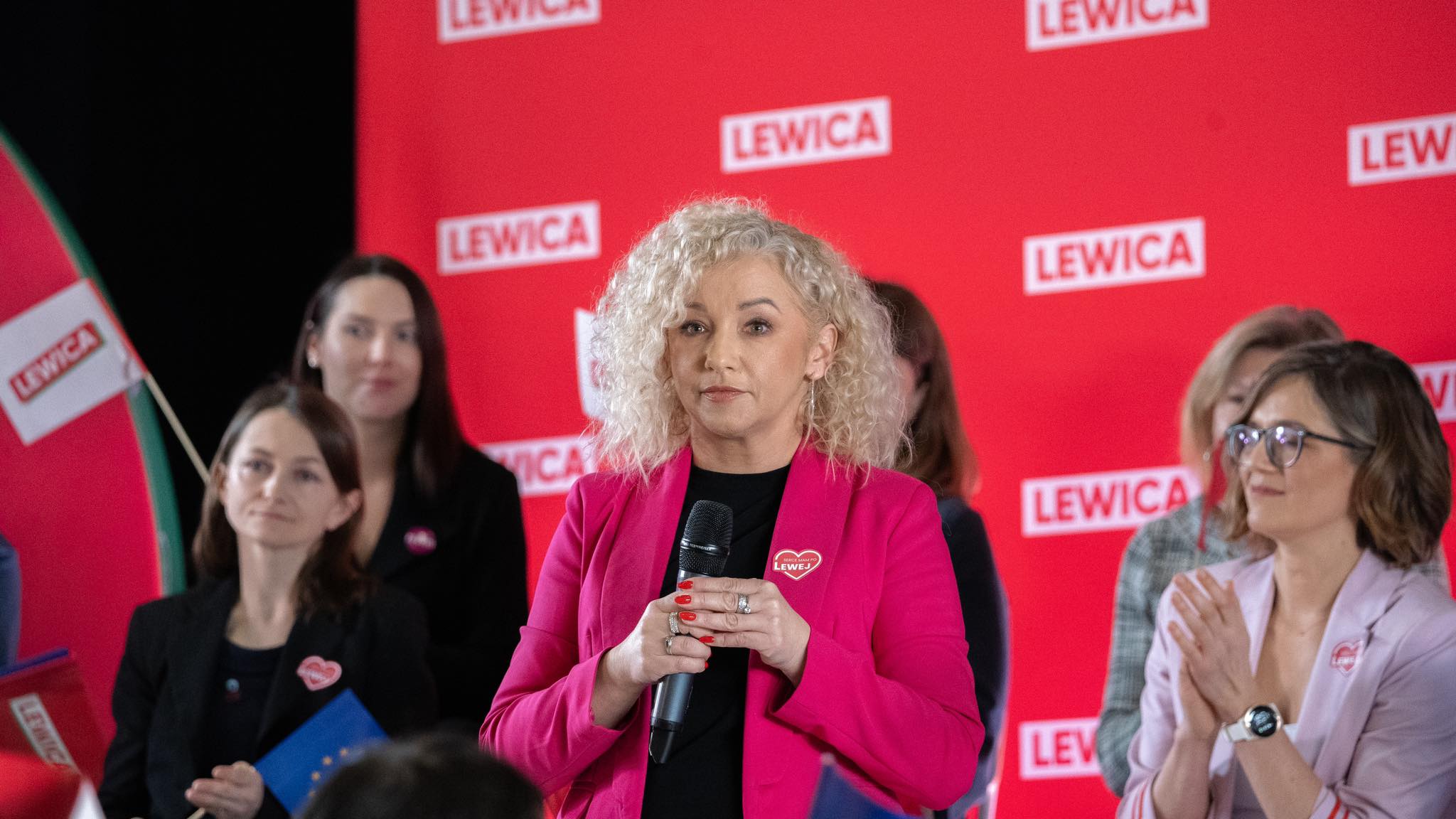 Kobieta z kręconymi blond włosami w różowym żakiecie przemawia do publiczności, trzymając mikrofon podczas wydarzenia Lewicy; w tle czerwone tło z logo LEWICA i inne osoby.