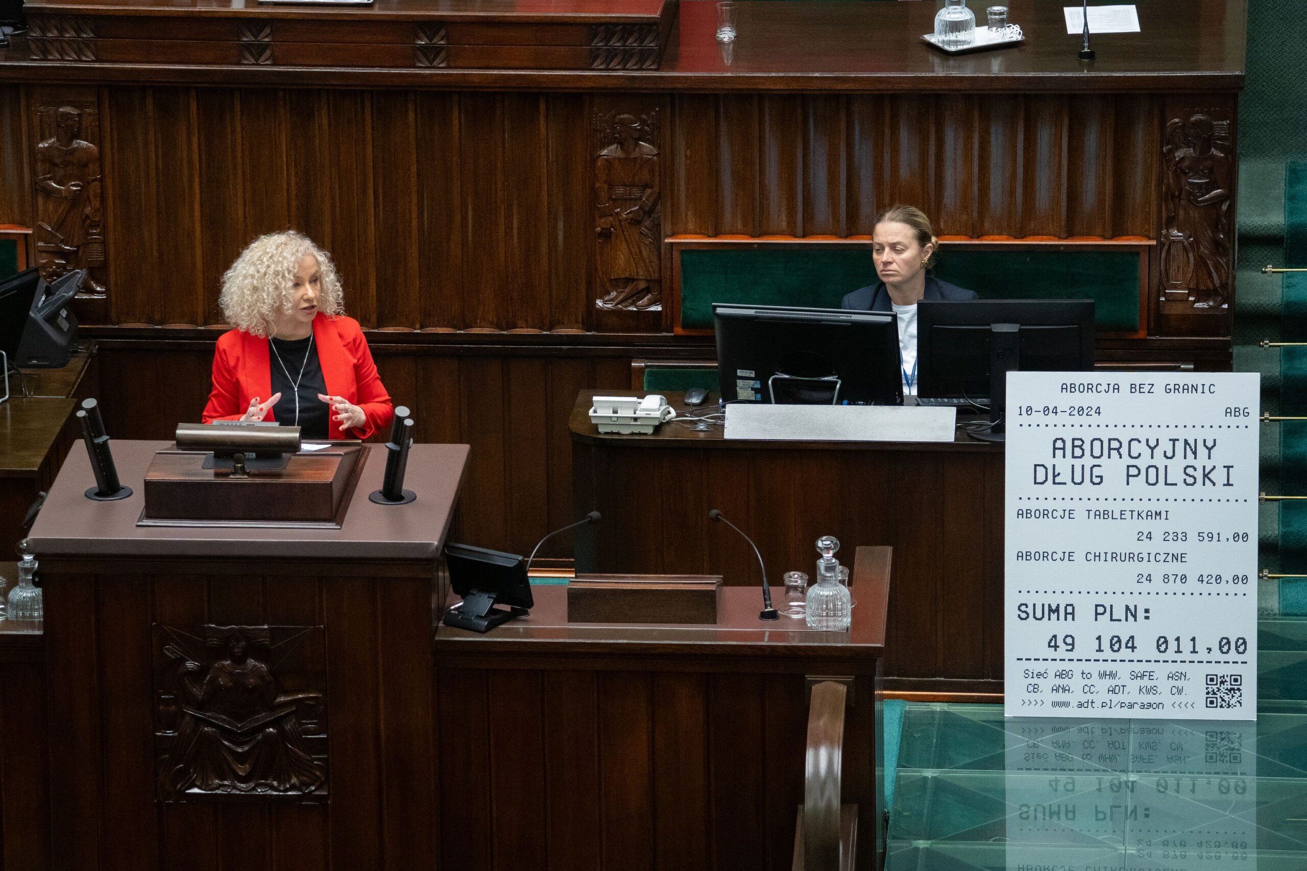 Kobieta w czerwonej marynarce wygłasza przemówienie z mównicy w sejmowej sali podczas debaty o aborcji, a po prawej stoi duży plakat przypominający rachunek z napisem „Aborcja bez granic”.