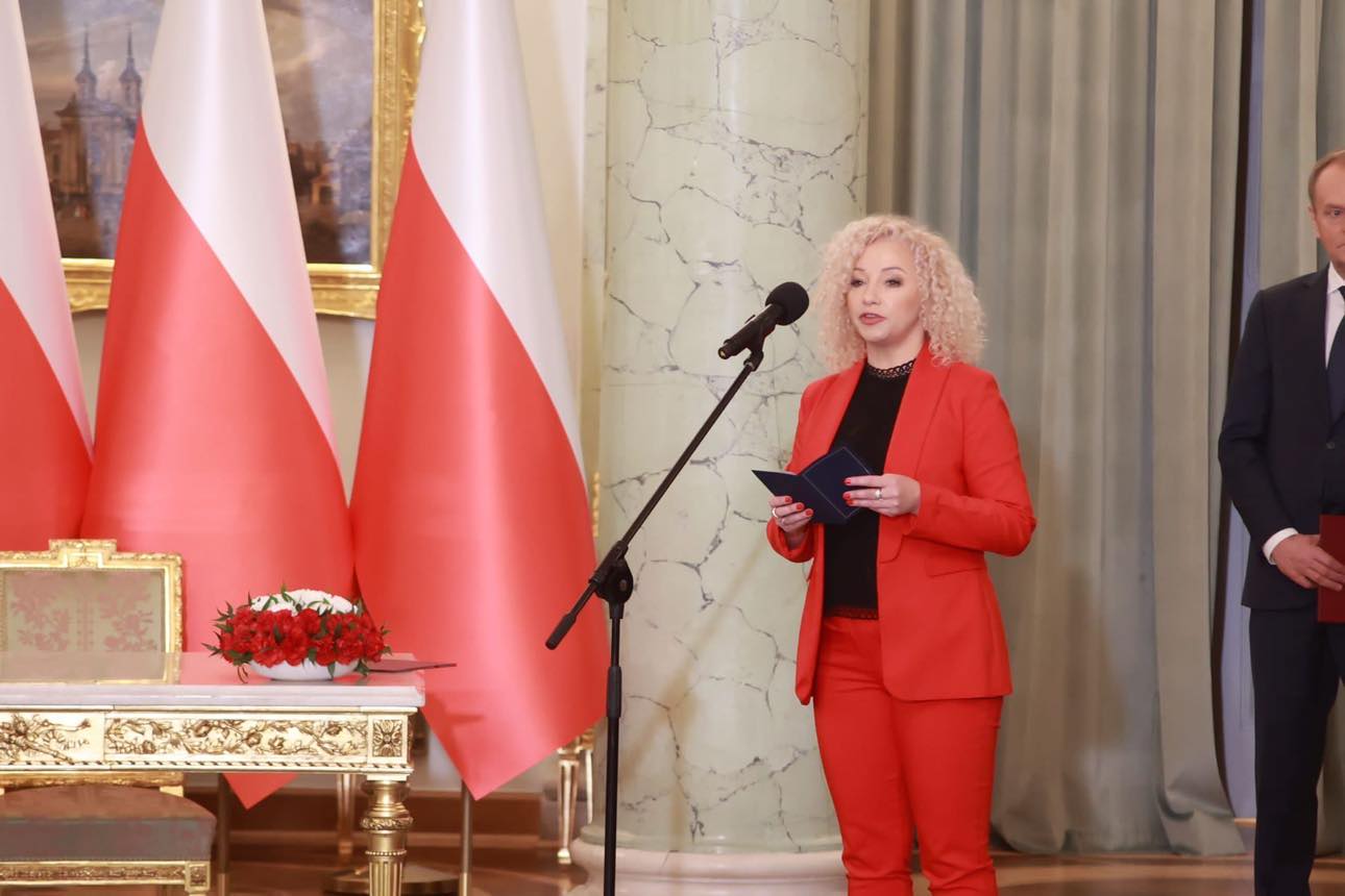 Kobieta w czerwonym garniturze przemawia do mikrofonu podczas zaprzysiężenia rządu, trzymając niebieski notes; w tle widoczne flagi Polski i marmurowa kolumna.