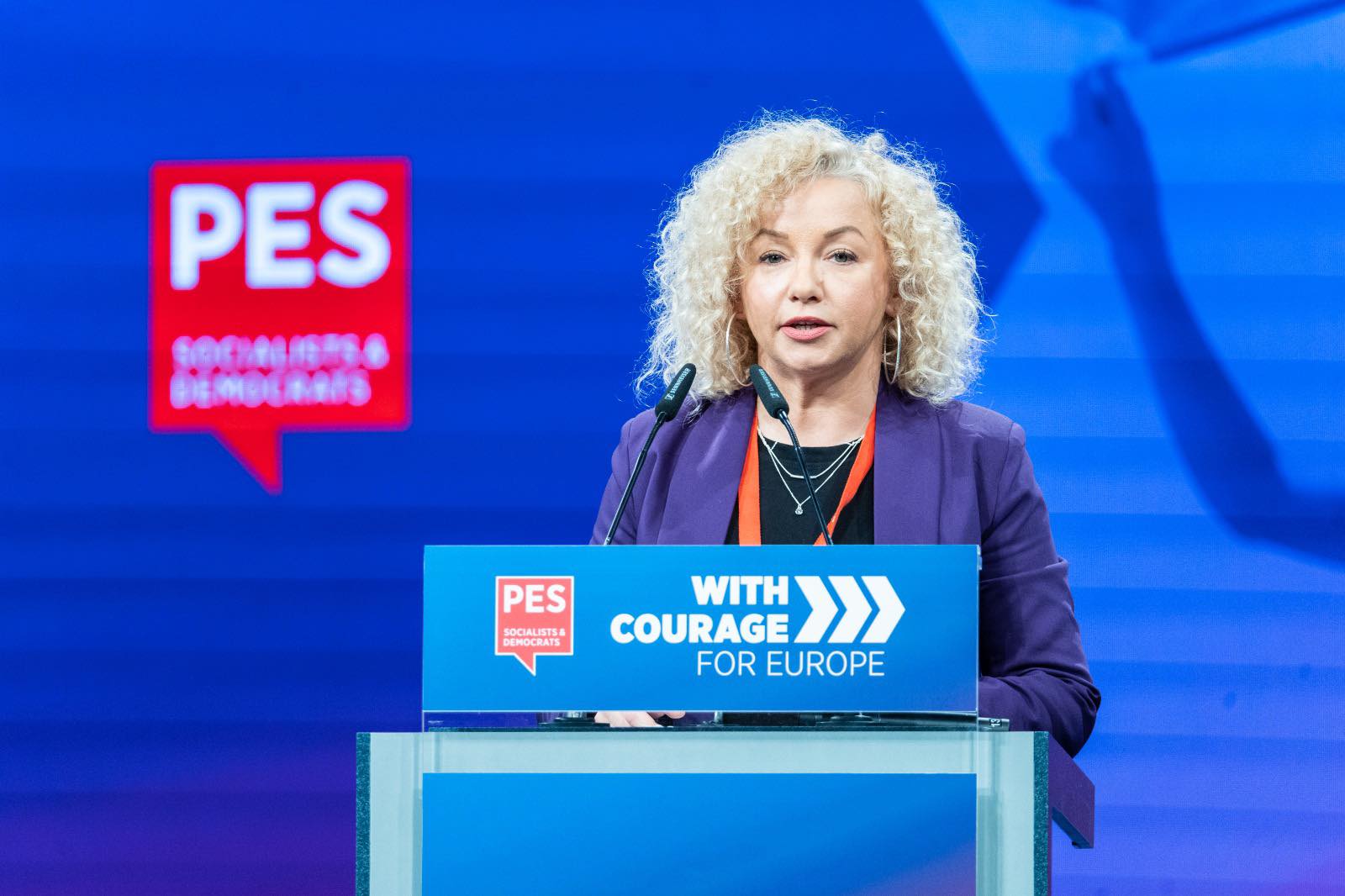 Na zdjęciu widać kobietę przemawiającą za mównicą podczas konferencji, z logo PES w tle.
