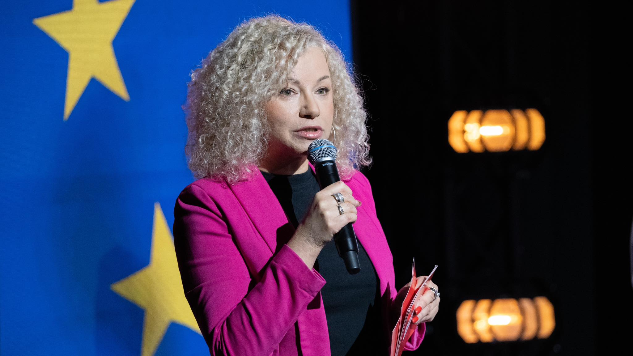 Kobieta z kręconymi blond włosami w różowej marynarce przemawia do mikrofonu podczas prezentacji programu wyborczego „Europa dla Ciebie”, na tle flagi Unii Europejskiej.