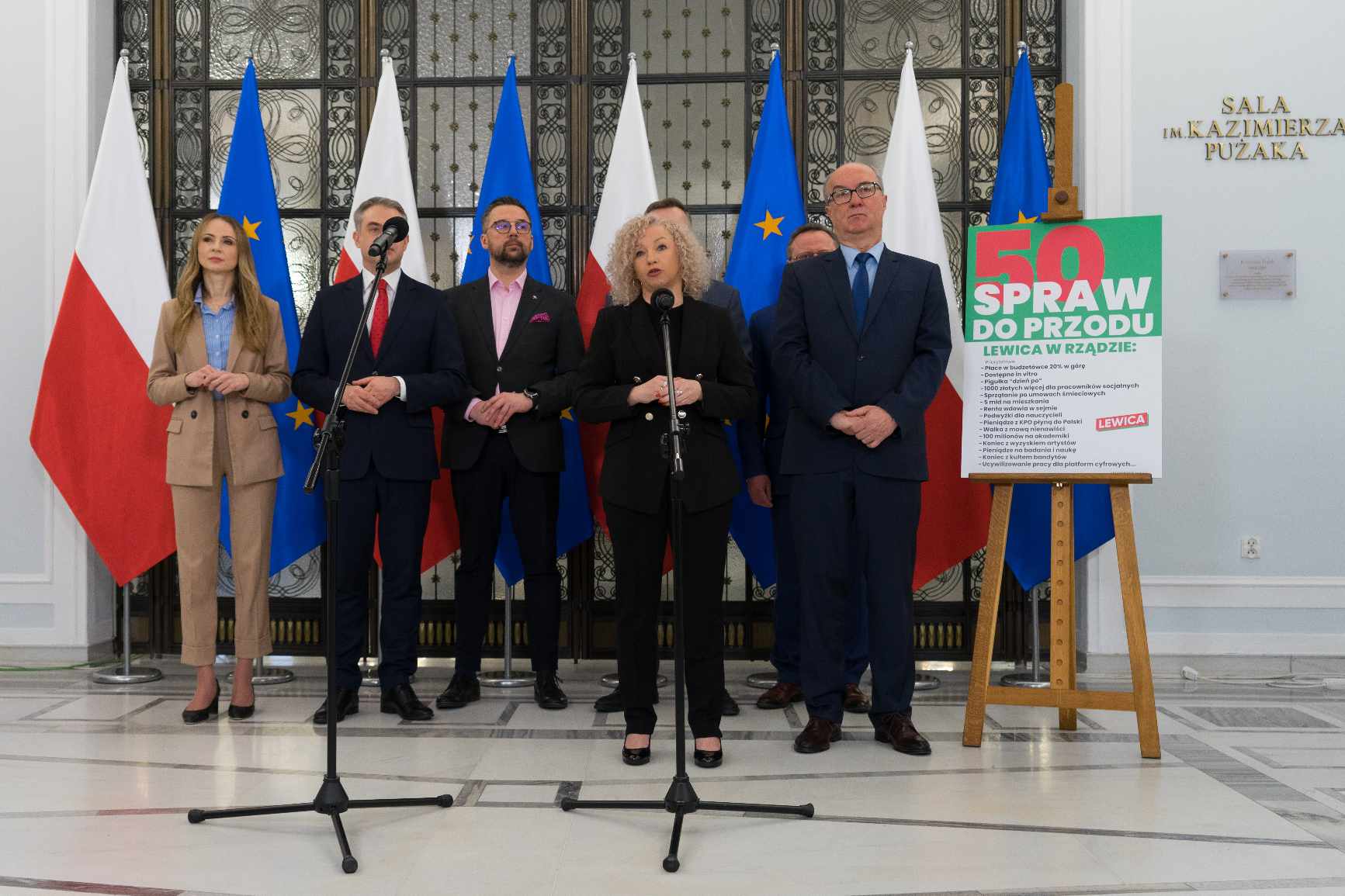 Grupa polityków w formalnych strojach stoi przed flagami Polski i Unii Europejskiej, obok plakatu z napisem „50 spraw do przodu”.