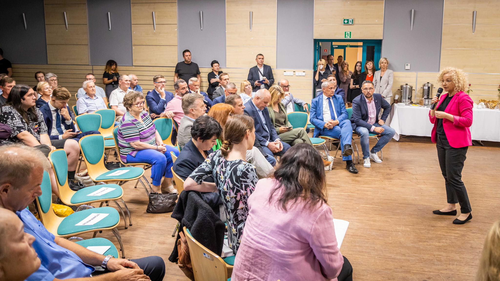 Grupa mieszkańców siedzi w sali konferencyjnej i słucha kobiety w różowym żakiecie, która przemawia do mikrofonu.
