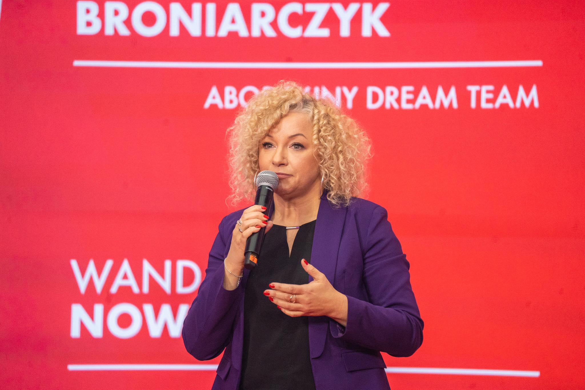 Kobieta z kręconymi włosami w purpurowej marynarce trzyma mikrofon i mówi na tle czerwonego tła z napisami.