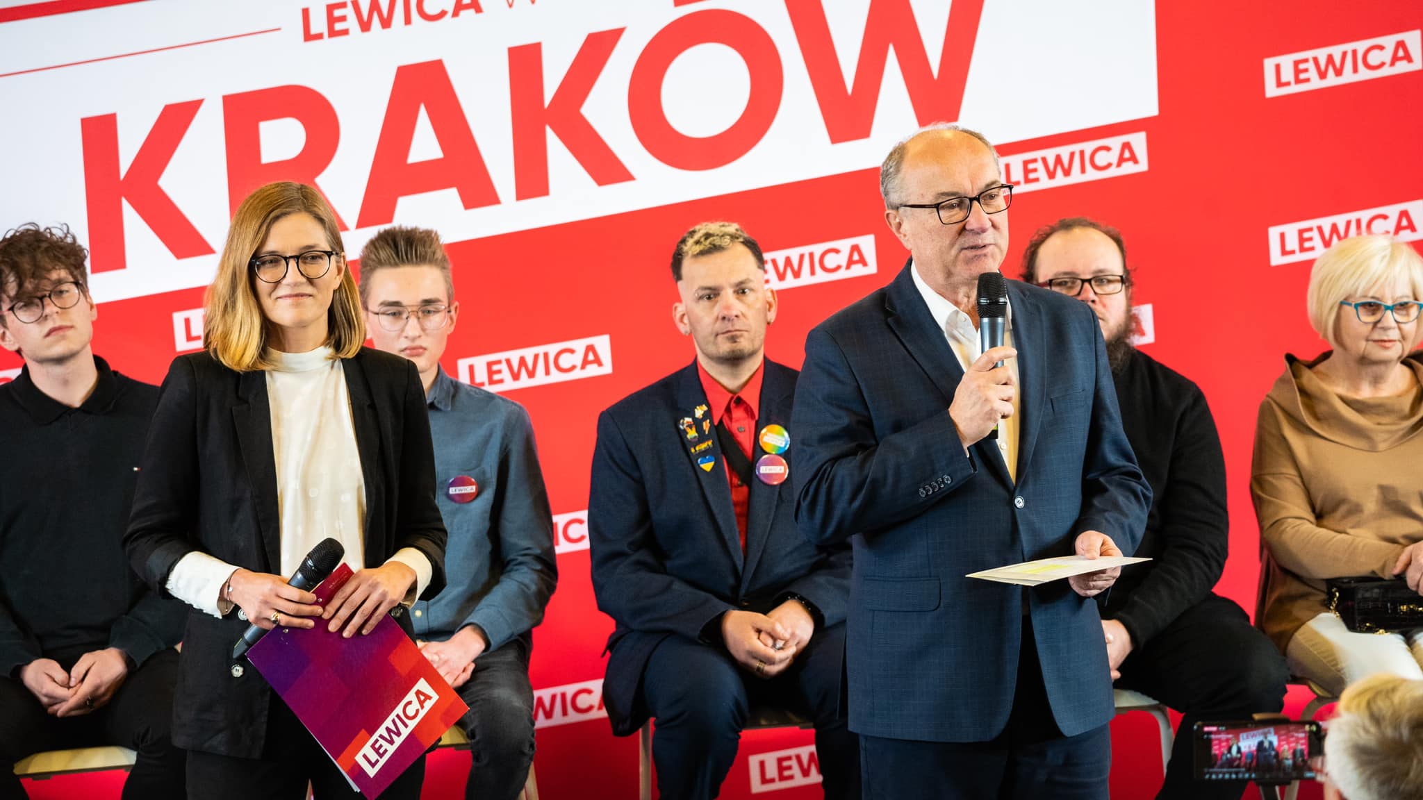 Grupa osób na scenie w Krakowie podczas wydarzenia Lewicy; kobieta w czarnym garniturze trzyma mikrofon i teczkę z logo Lewicy, podczas gdy mężczyzna w garniturze przemawia do mikrofonu, a w tle widać czerwone tło z napisem KRAKÓW i logotypami Lewicy.