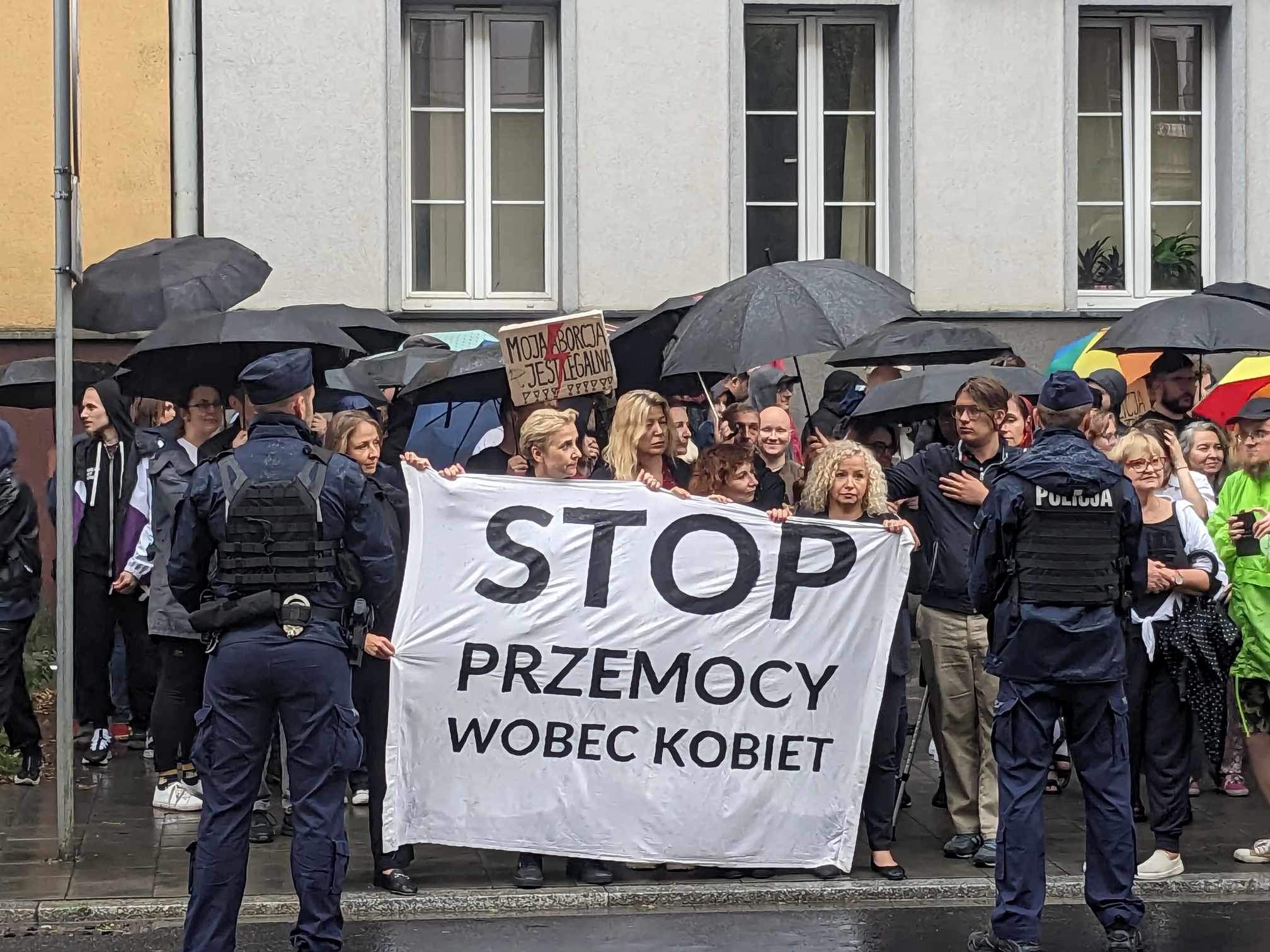 Grupa protestujących w Krakowie trzyma biały baner z napisem STOP PRZEMOCY WOBEC KOBIET podczas deszczowej pogody, na pierwszym planie stoją policjanci.