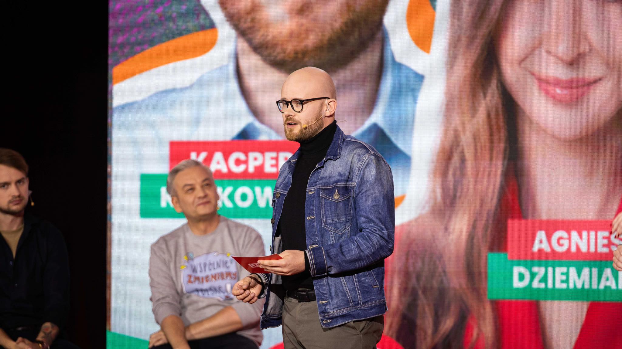 Mężczyzna z okularami i krótką brodą, ubrany w czarny golf i denimową kurtkę, prowadzi wystąpienie na scenie z mikrofonem, trzymając kartkę; w tle widać duży ekran z wizerunkami młodych osób i kolorowymi napisami.