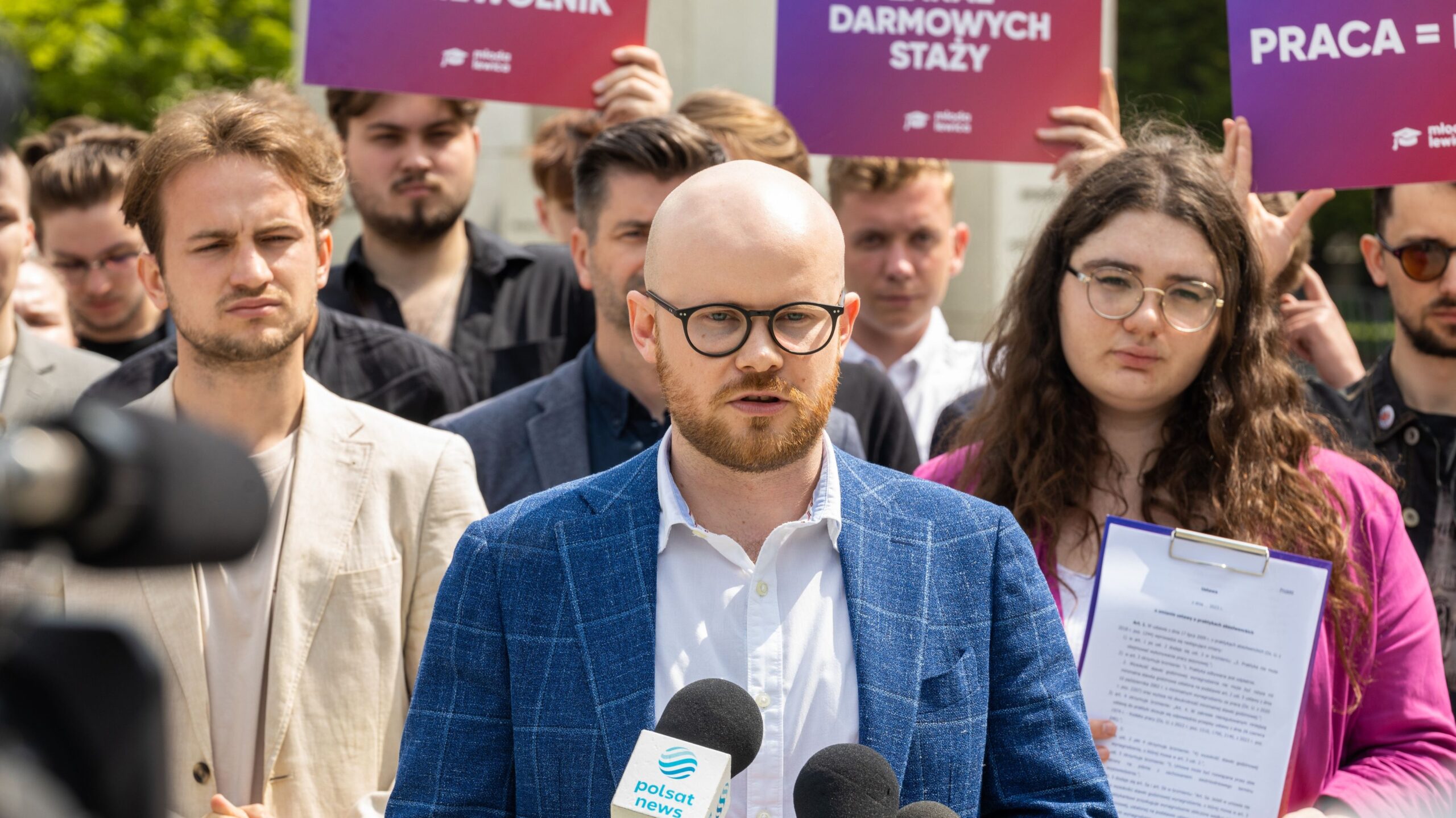 Mężczyzna z okularami i brodą w granatowej marynarce przemawia do mikrofonów podczas protestu o wynagrodzenie za pracę, wokół niego demonstranci trzymają plakaty „PRACA = PŁACA”.